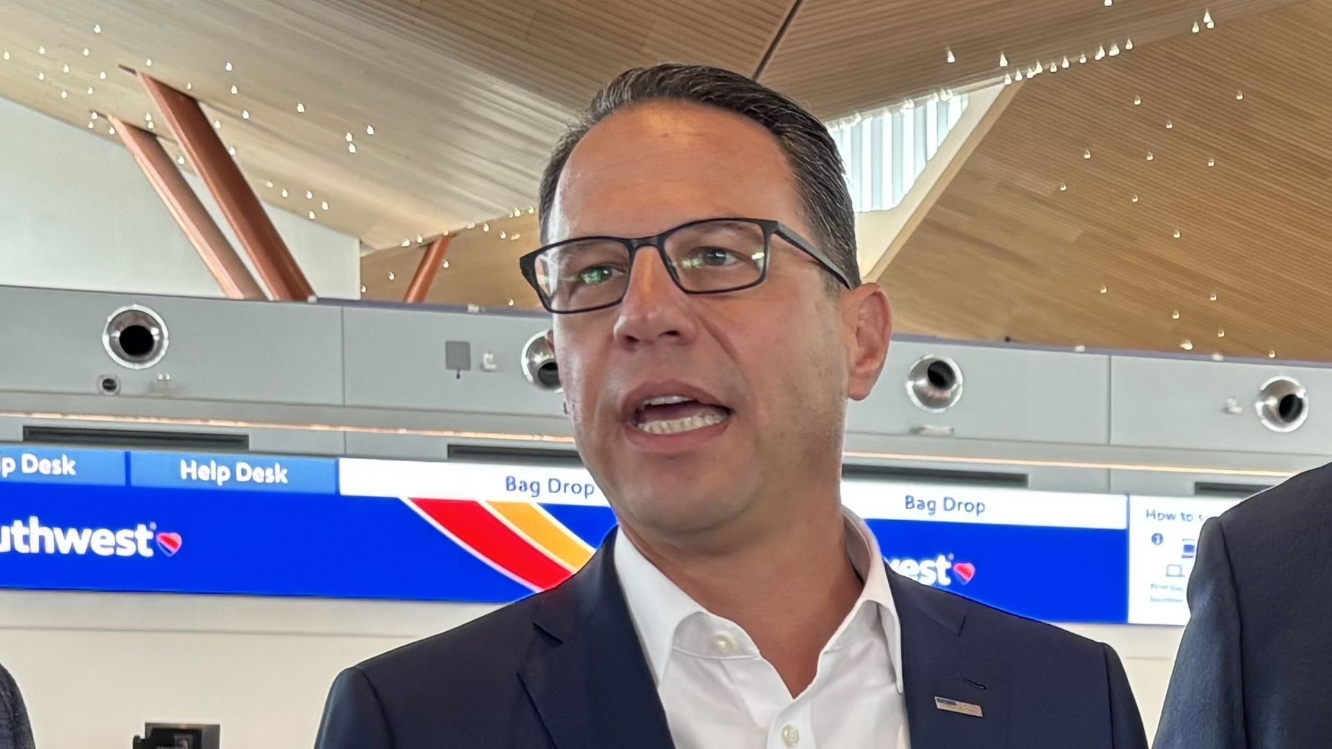 Man in a navy suit and white shirt speaking indoors, wearing black-rimmed glasses, with a blue Southwest Airlines help desk sign in the background inside an airport terminal.