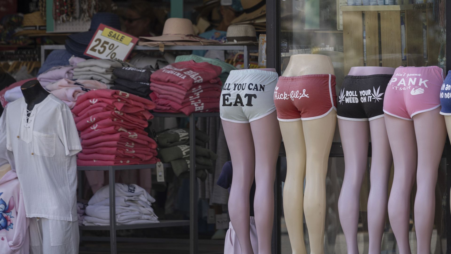 Store display with mannequins wearing colorful shorts with humorous phrases, stacks of folded clothes, hats, and a 25% off sale sign inside the shop.