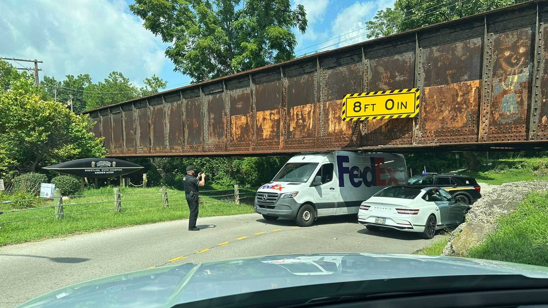 Rusted steel railroad bridge spans a road beneath a blue sky. A bright yellow clearance sign reads 8 FT 0 IN. A FedEx van and a man in dark clothing stand near green trees.