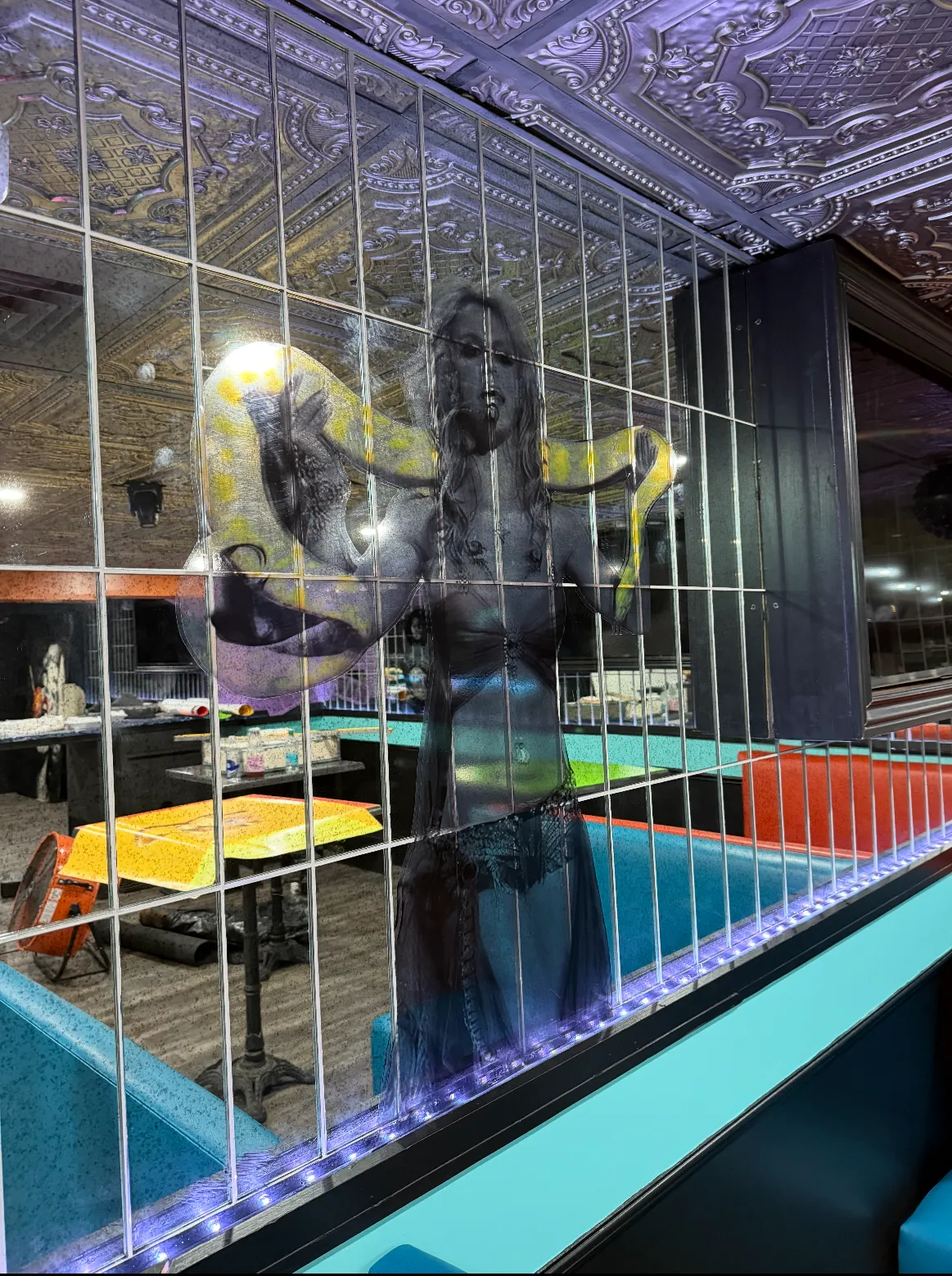 Reflection of a woman with a yellow and white python draped over her shoulders on a mirrored wall, inside a restaurant with turquoise and red booths and decorative ceiling tiles.