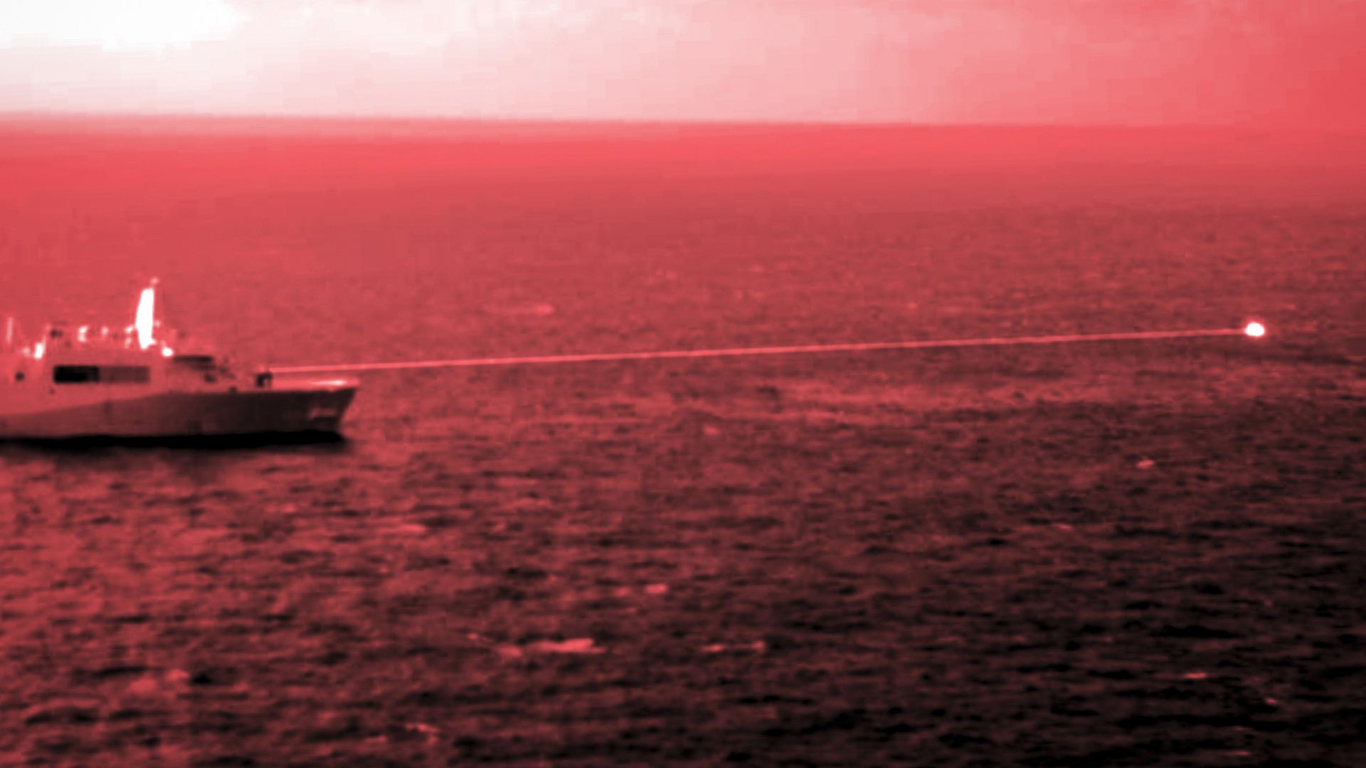 Ship on ocean firing a visible bright laser beam across the water under a cloudy sky, with red tint over the image.