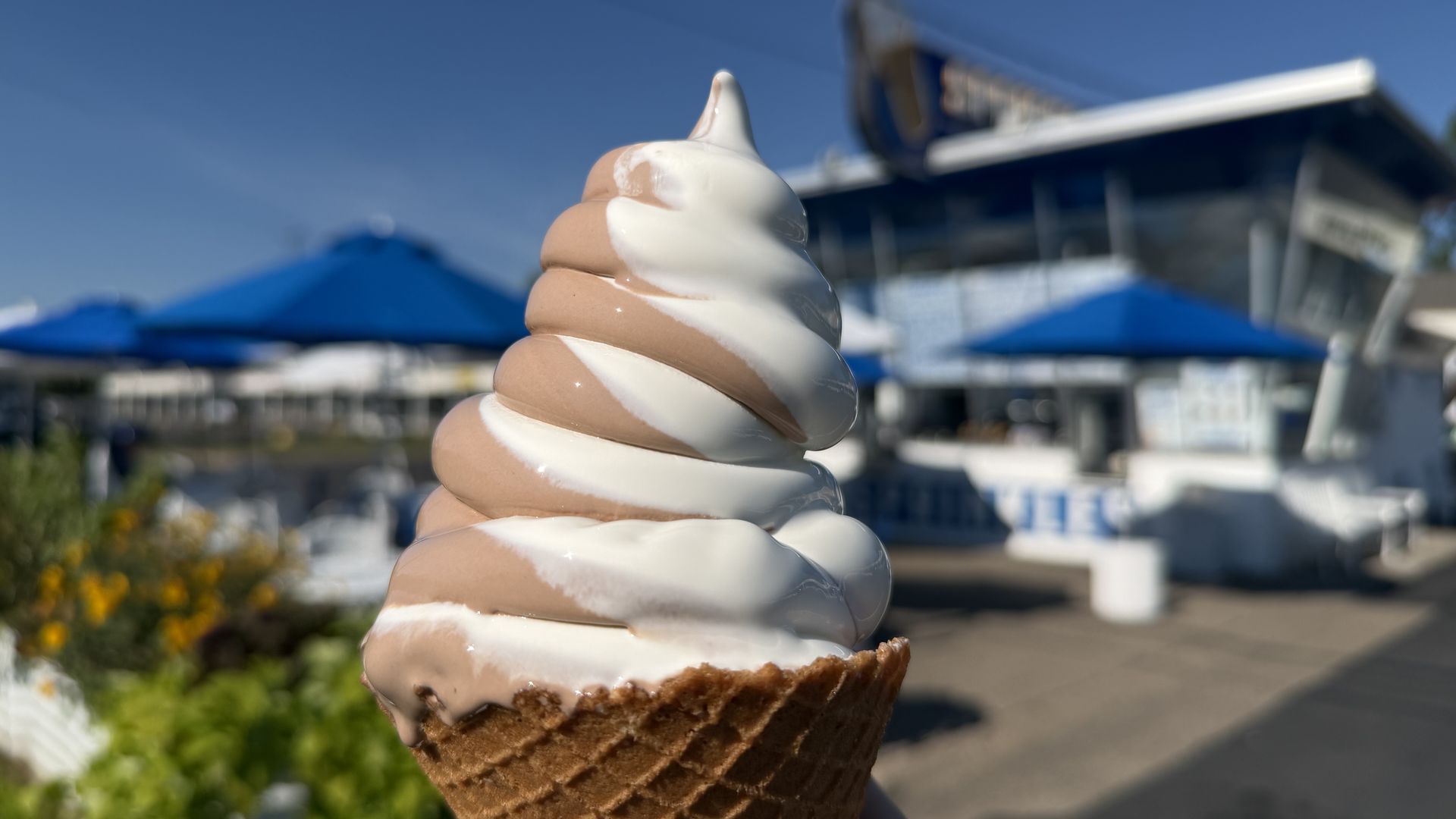 Close-up of a vanilla and chocolate swirled soft-serve ice cream in a waffle cone, held up against a blue sky with blurred outdoor umbrellas and a modern building in the background.