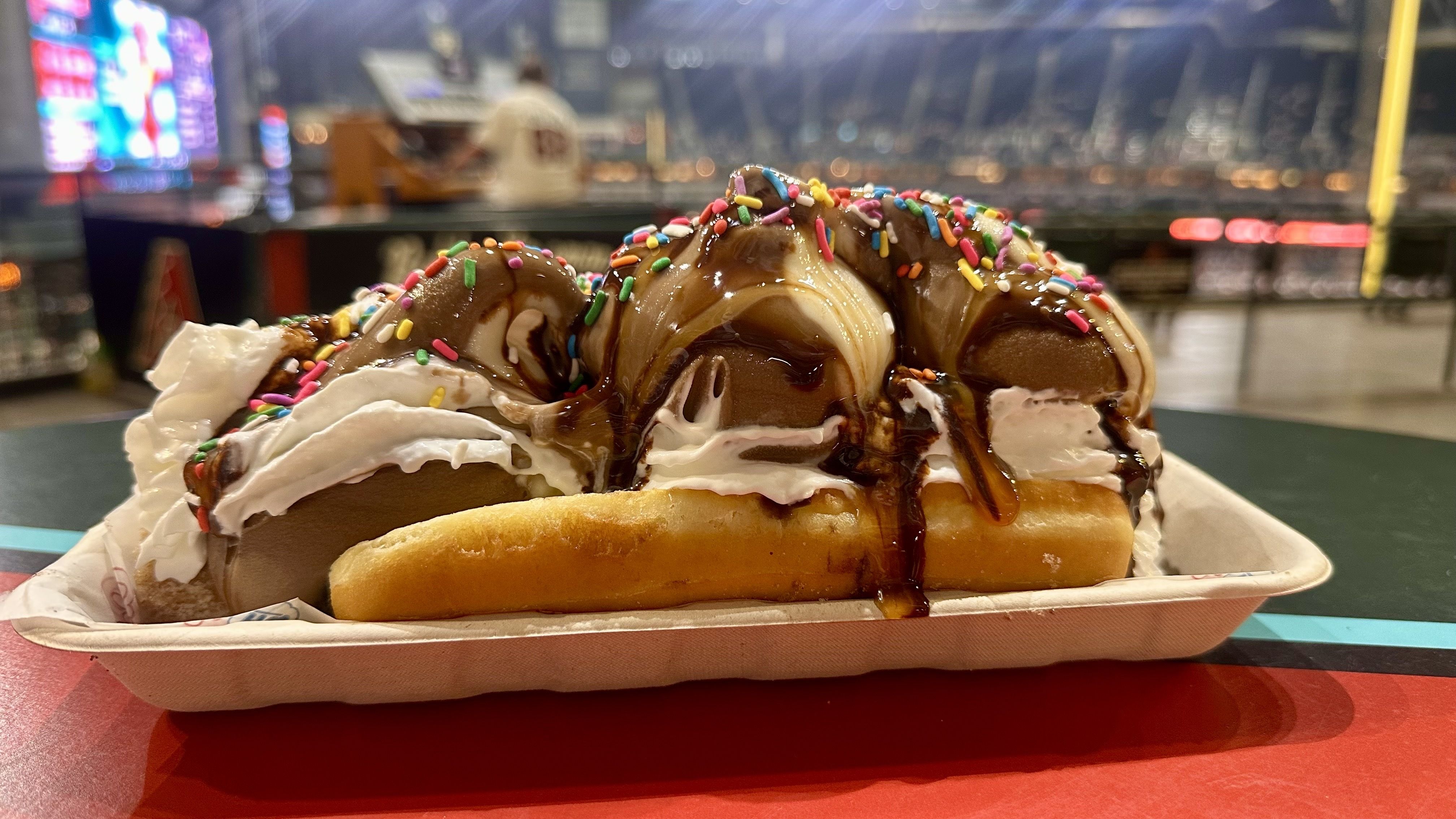 Ice cream sundae with chocolate and vanilla scoops, whipped cream, chocolate syrup, and colorful sprinkles on a tray, at a well-lit stadium or event venue in the evening.