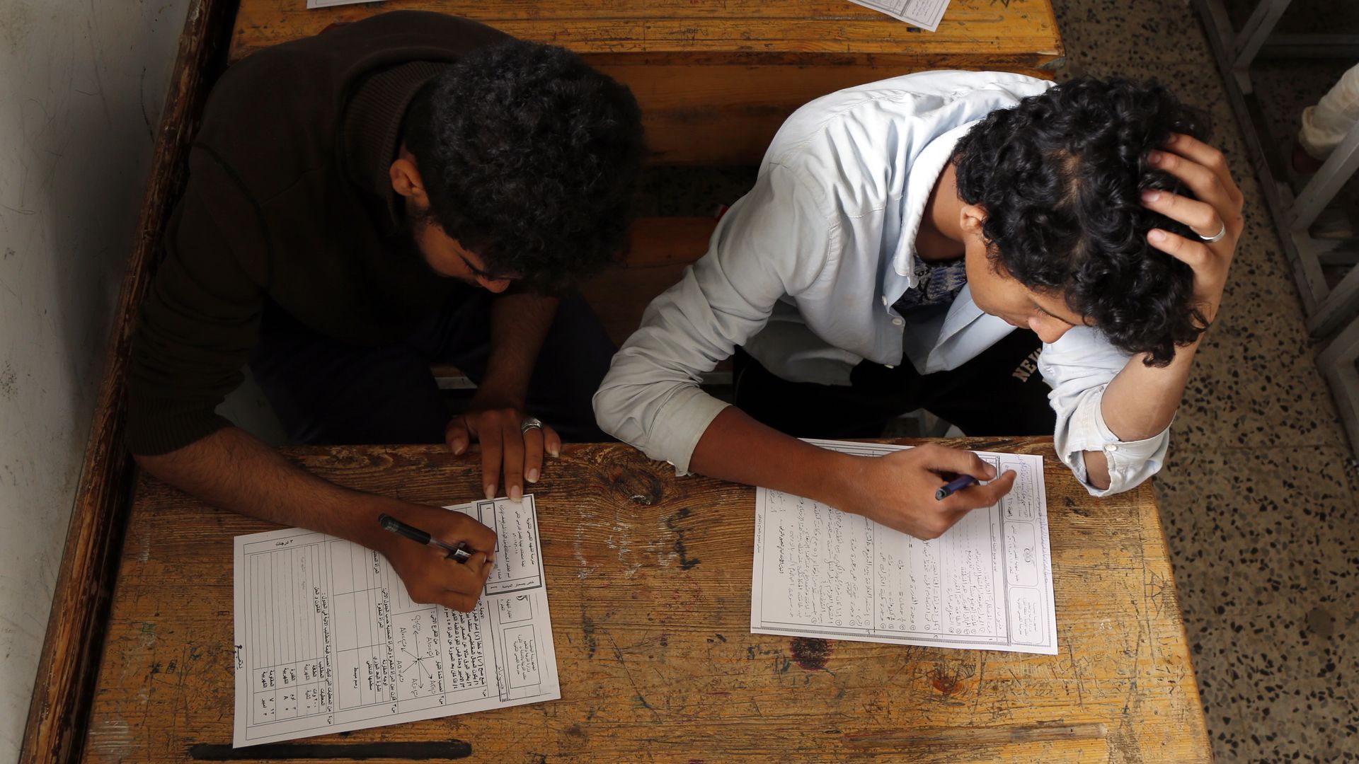 In this image, two male students sit at a desk while taking a test on separate sheets of paper.