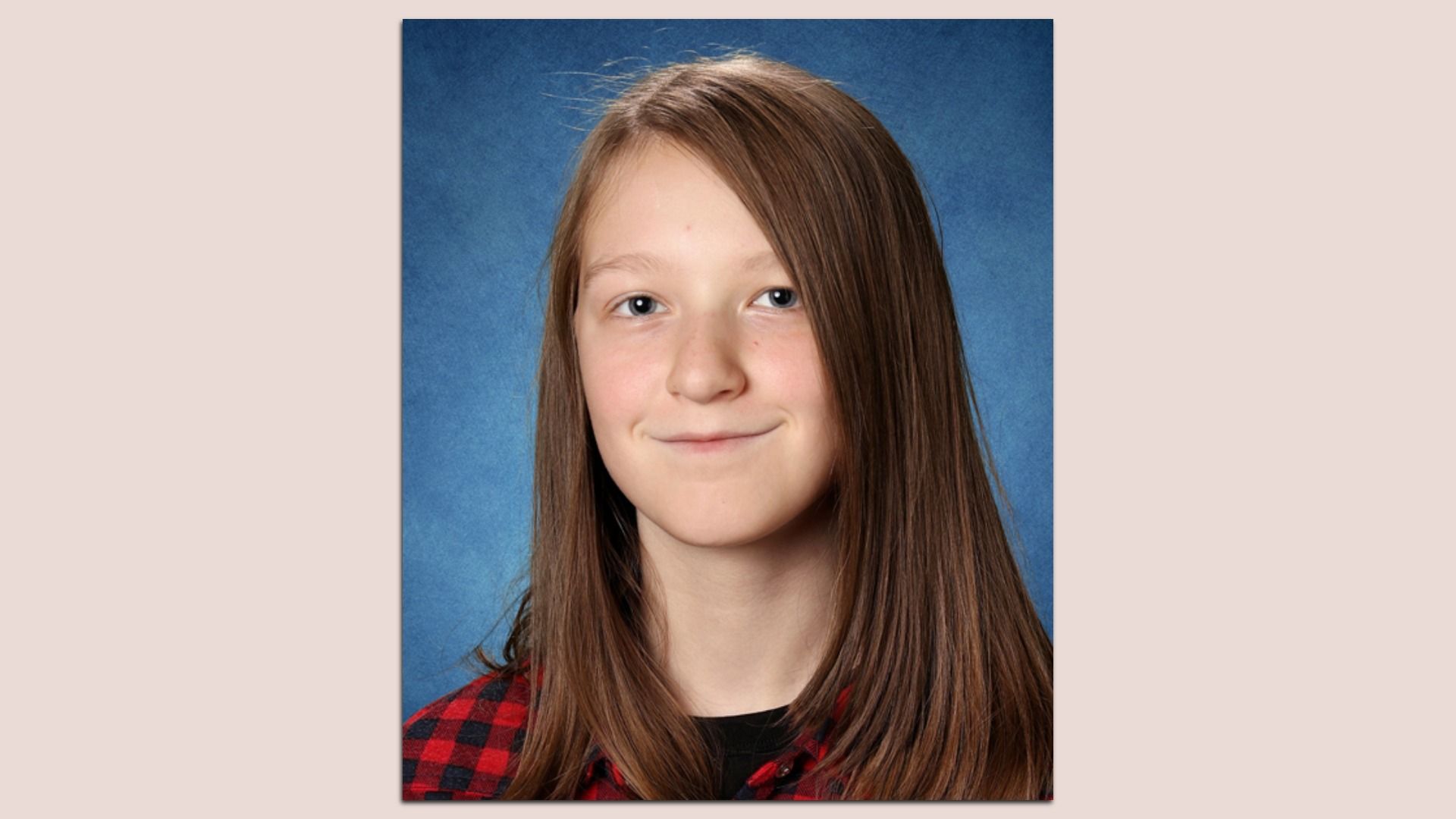 Portrait of a smiling teenage girl with long brown hair, wearing a red and black plaid shirt, against a blue studio backdrop.