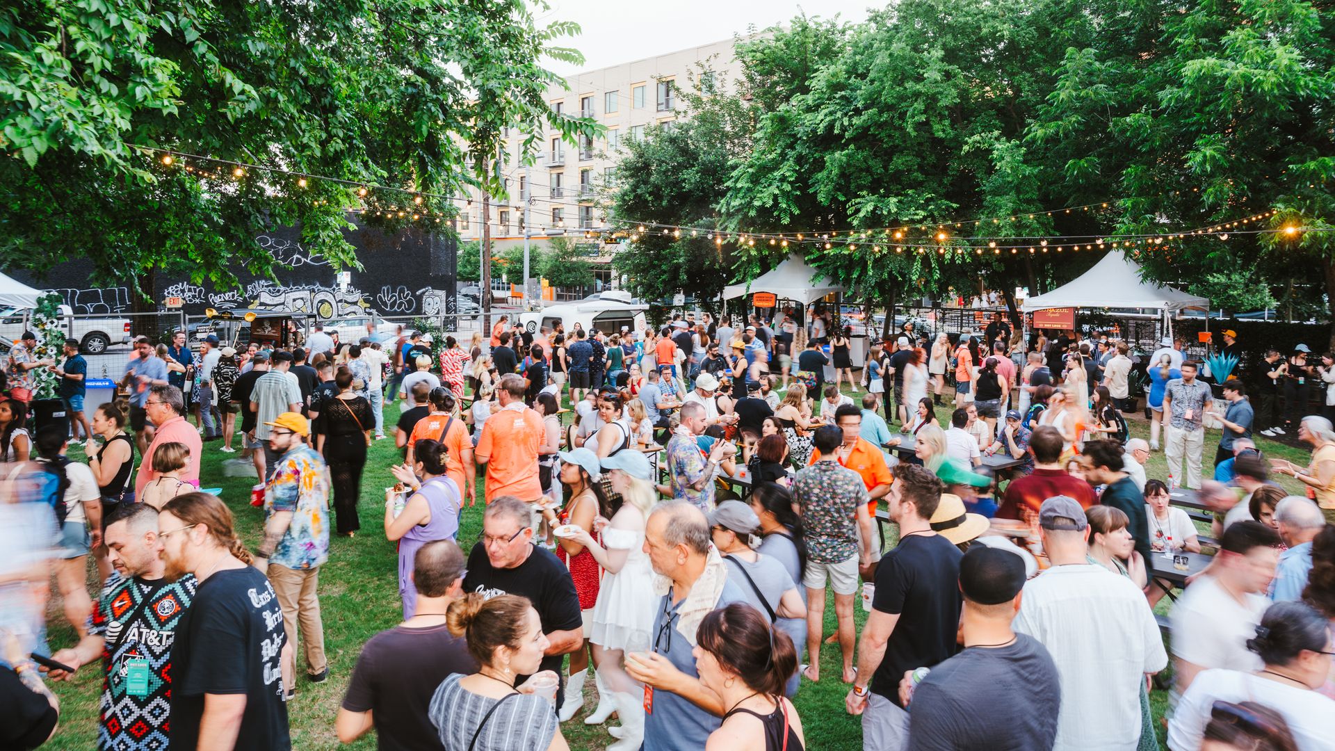 Crowded outdoor festival in a city park with string lights, lush trees, white tents, and a graffiti-covered wall; people mingle, chat, and enjoy drinks on the grass.