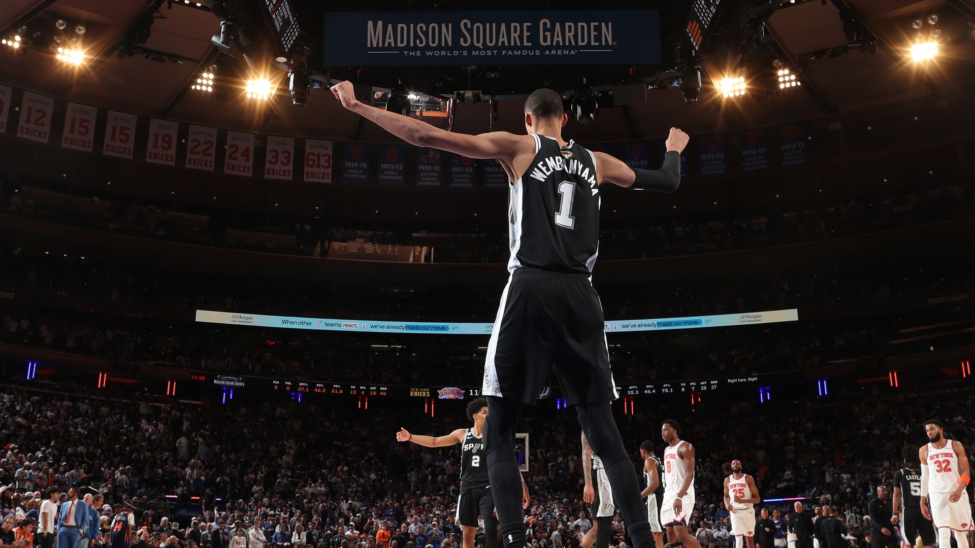 Basketball player wearing jersey number 1 with name Wembanyama in a black uniform raises his arms on the court at Madison Square Garden; bright arena lights, banners and a crowd in the background.