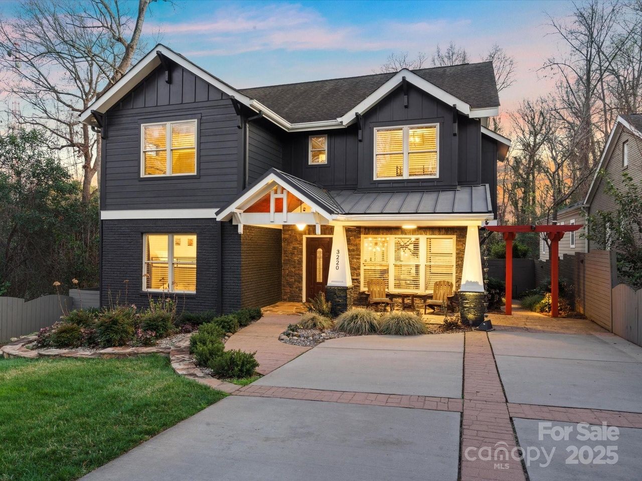 Two-story dark gray house with white trim and warm interior lights, a stone pathway, front porch with two wooden chairs, landscaped garden, and concrete driveway at dusk.