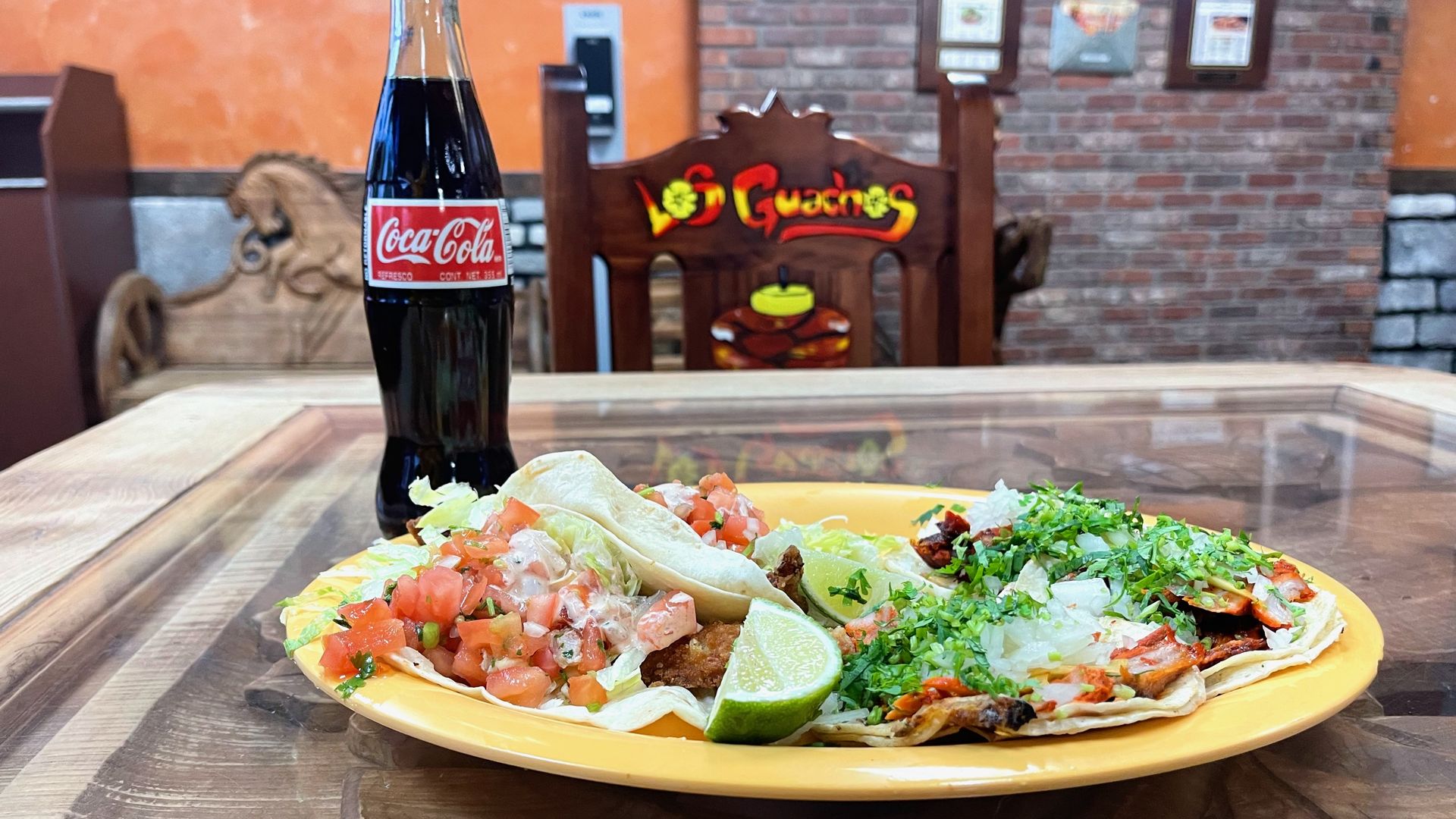 A yellow plate of tacos with a glass Coke bottle next to it and a chair in the background that reads "Los Guachos"