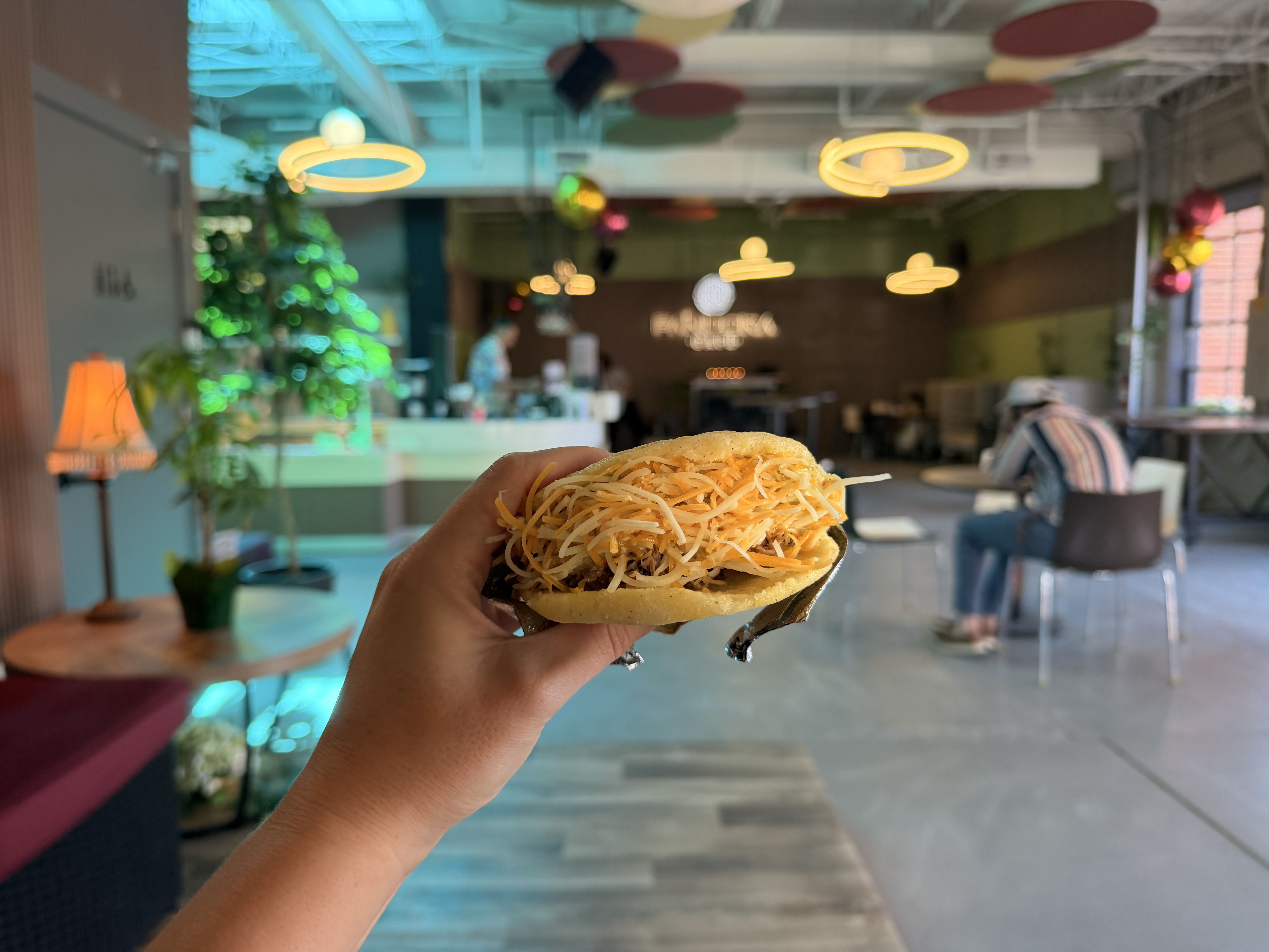 A hand holds a small sandwich with shredded cheese filling inside a soft bun, in a bright, colorful cafe with circular ring lights, plants, and customers in the background.