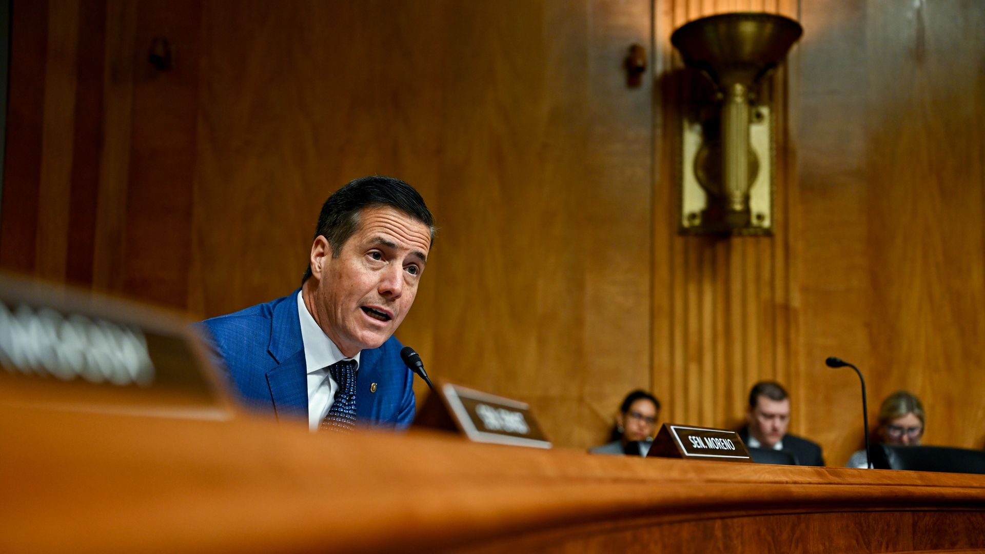 Man in blue suit in congressional hearing, wood paneled room, asking questions into microphone. 