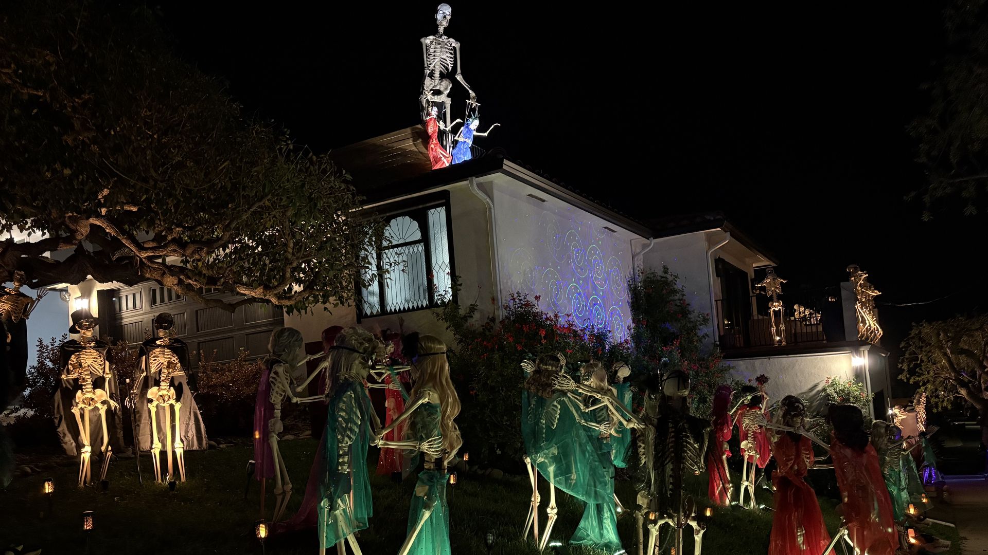 Night scene of a house decorated with numerous illuminated skeletons dressed in colorful cloaks, arranged in pairs and groups on the lawn and roof, with tree and pathway lights.