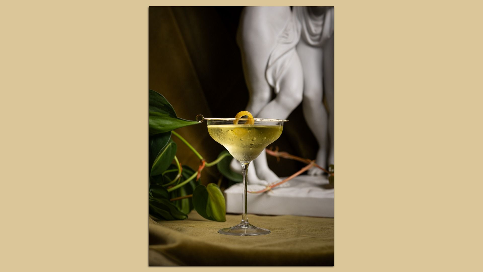 Clear cocktail in a delicate glass with lemon peel garnish on a skewer, set against a backdrop featuring green leaves and part of a white classical statue's legs.