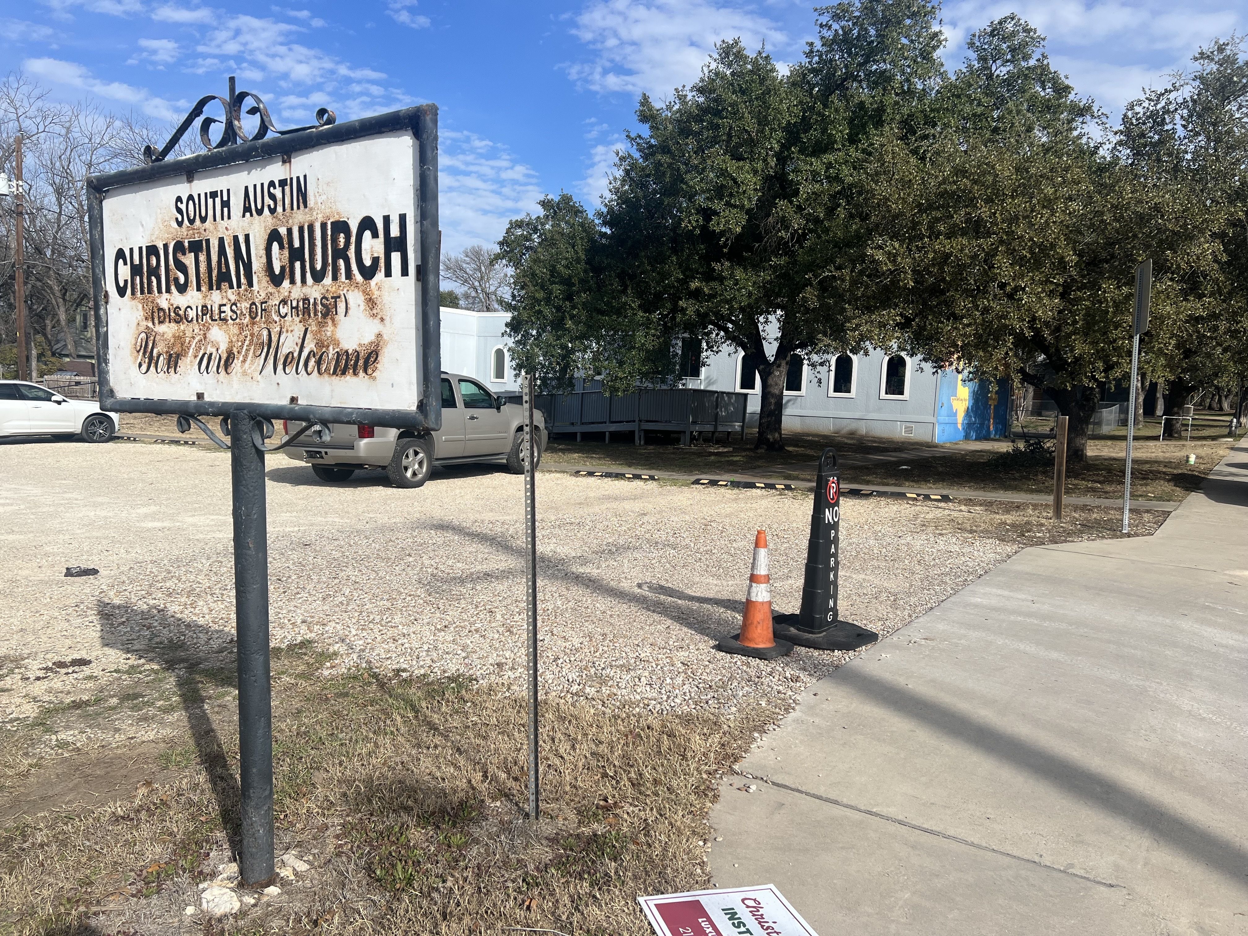 Rusty white sign reading "South Austin Christian Church (Disciples of Christ) You are Welcome" near gravel parking lot with cars, trees, and blue sky on a sunny day.