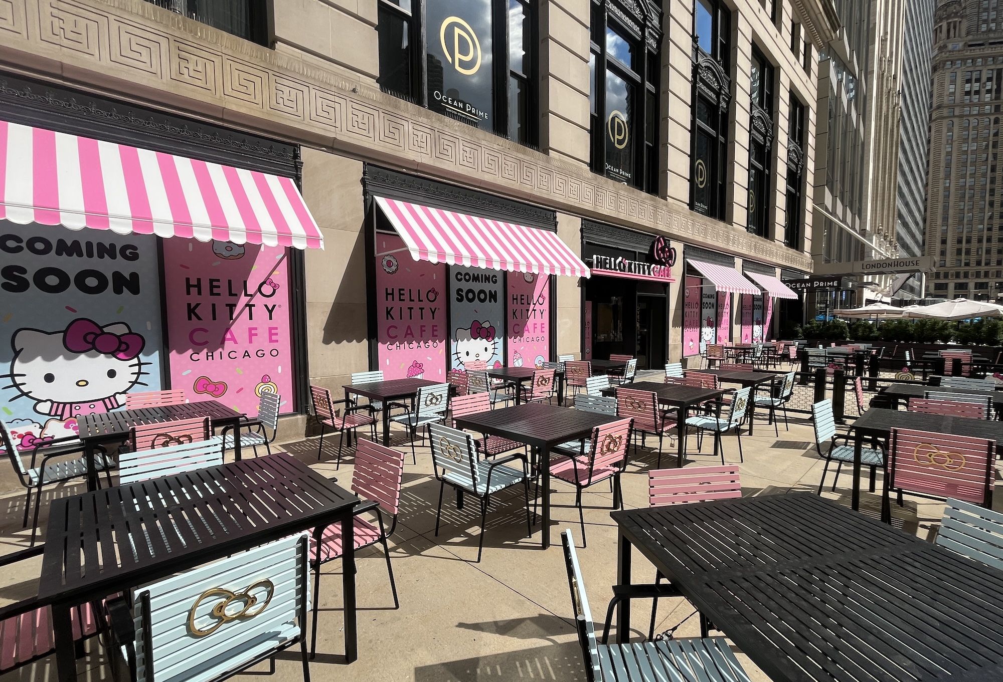 Wide sidewalk cafe with pink striped awnings and pastel blue chairs, Hello Kitty Cafe signage on pink panels, metal tables, in a sunny Chicago block with tall beige buildings and outdoor seating.
