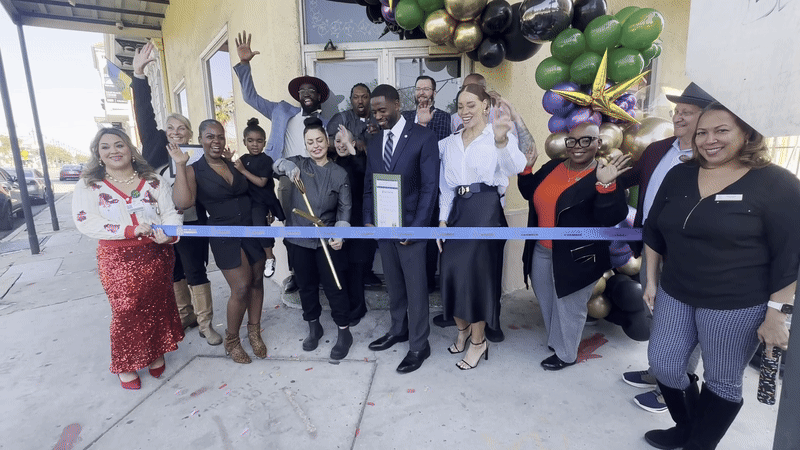 A group of people celebrate as a person in a chef's coat cuts a long blue ribbon in front of a doorway.