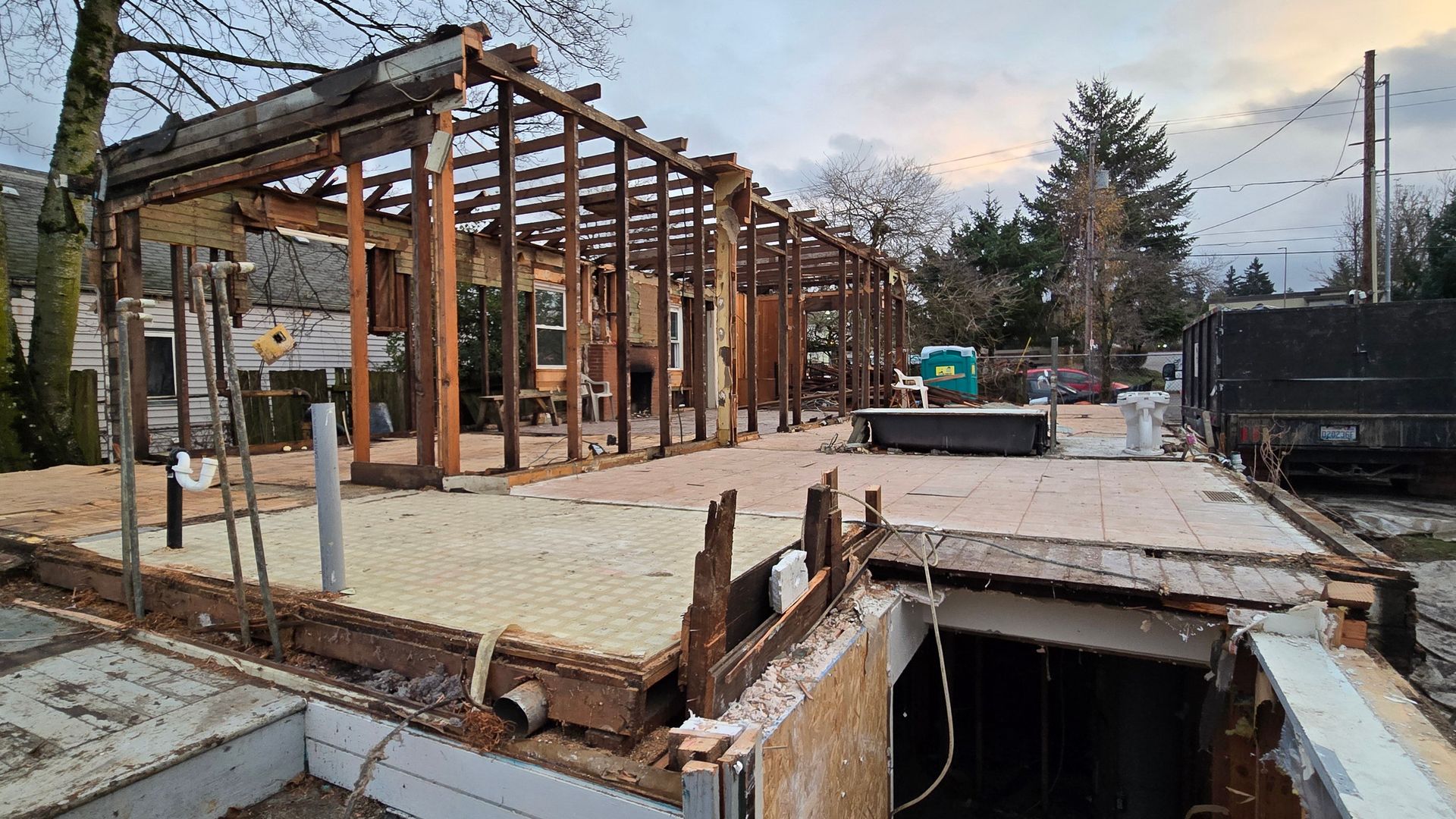 Backyard demolition: exposed wooden frame and rafters of an ongoing extension, with tiled floor, construction debris, a dumpster, a portable toilet, and an open foundation pit in the foreground.