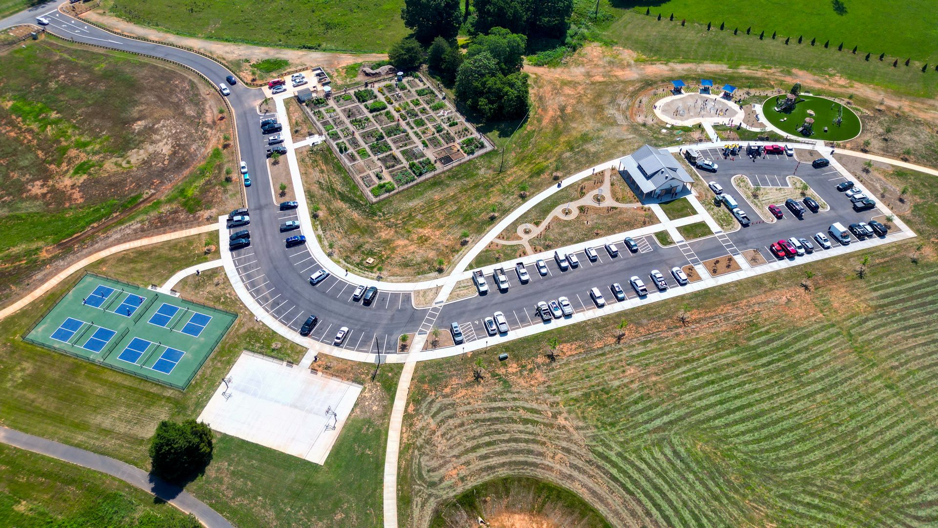 Aerial view of a park and campus: curved roads, a grid of garden beds, parking lots full of cars, a small building near circular space with playground equipment, and tennis courts at bottom left.