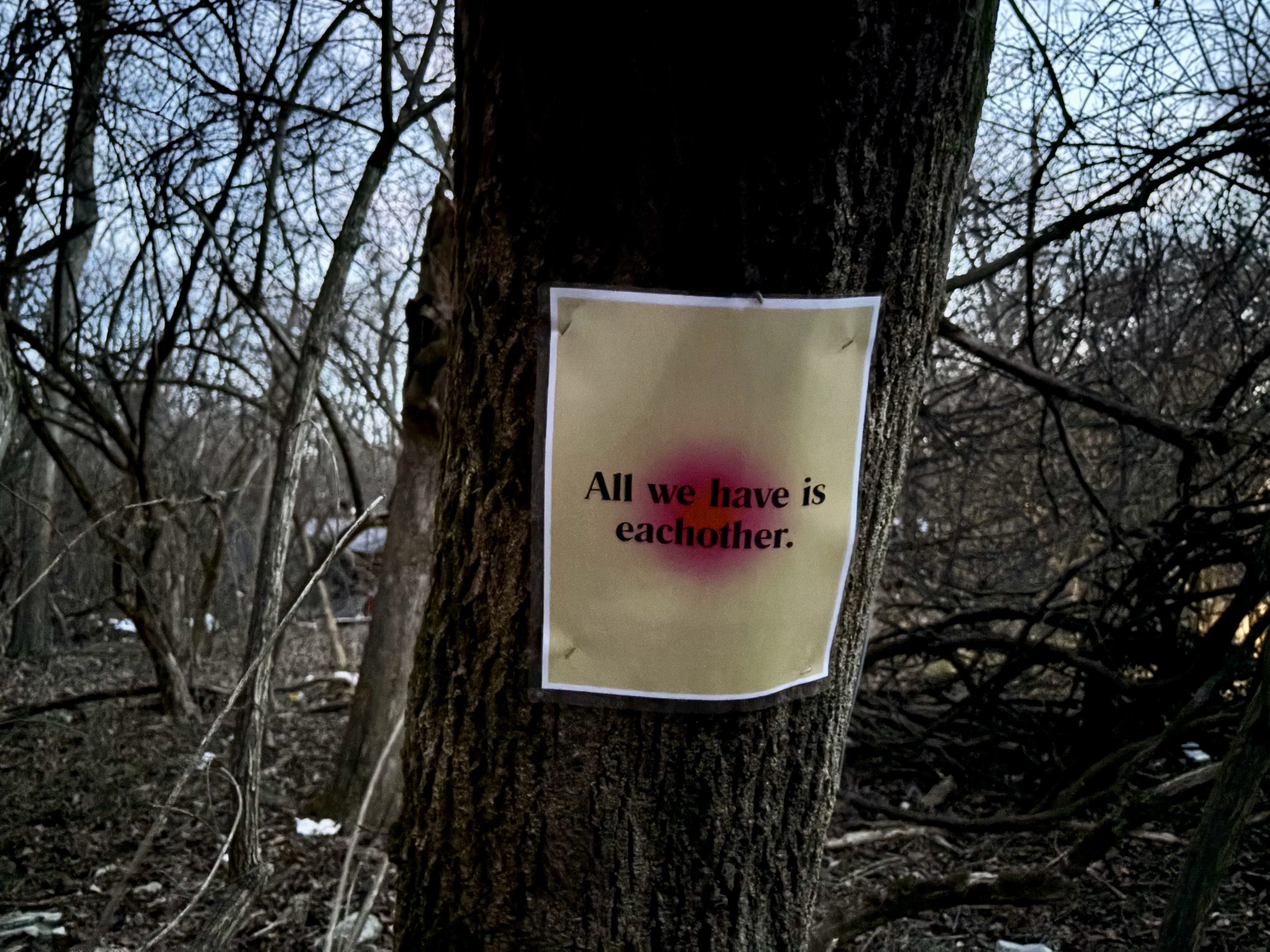 Paper sign with text "All we have is eachother." pinned to a large tree trunk in a leafless, wooded area under a fading sky.