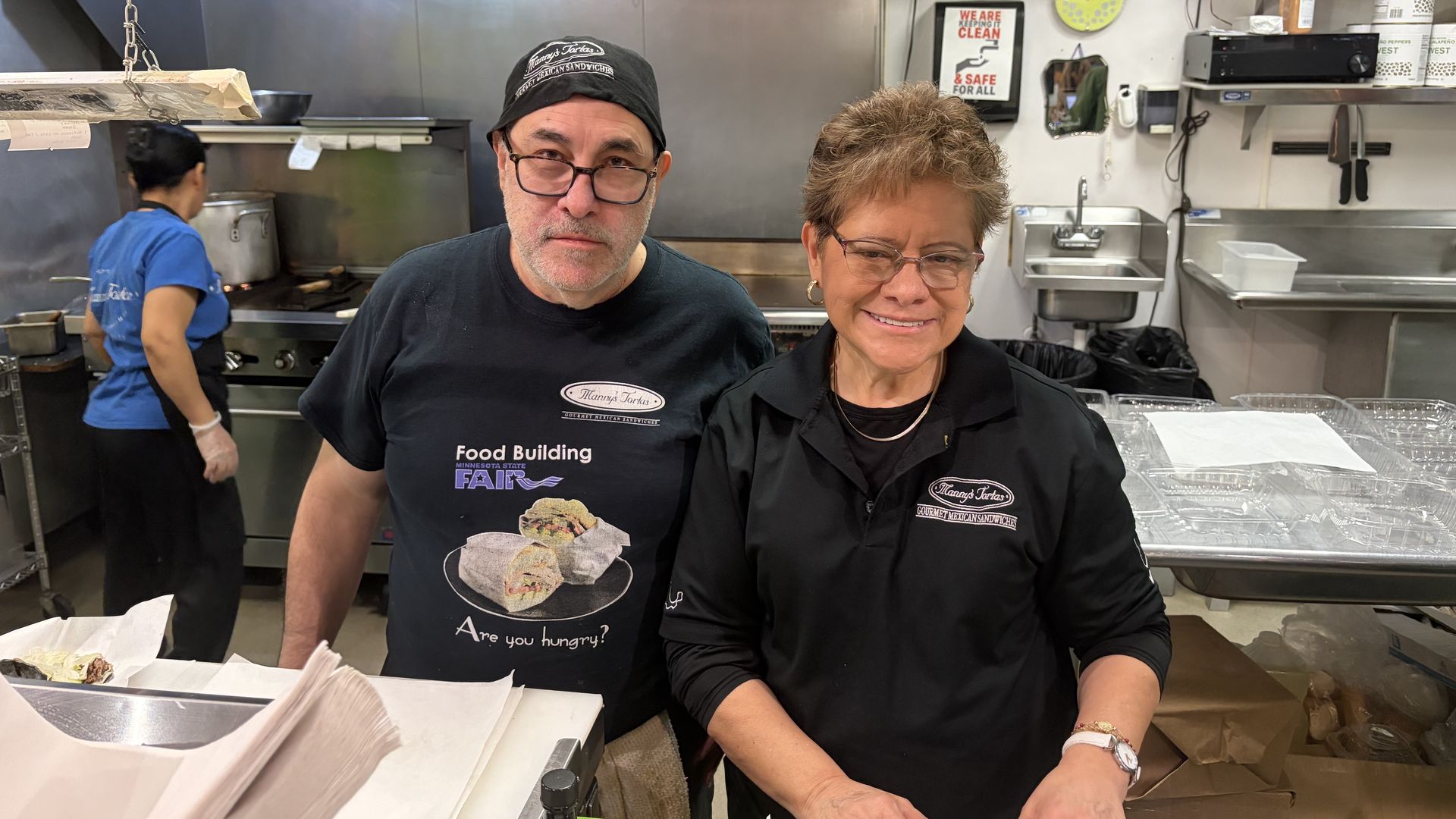 Two people in a kitchen wearing black shirts and glasses, standing behind a counter with food containers, with a third person cooking in the background. A sign above shows social media handles.