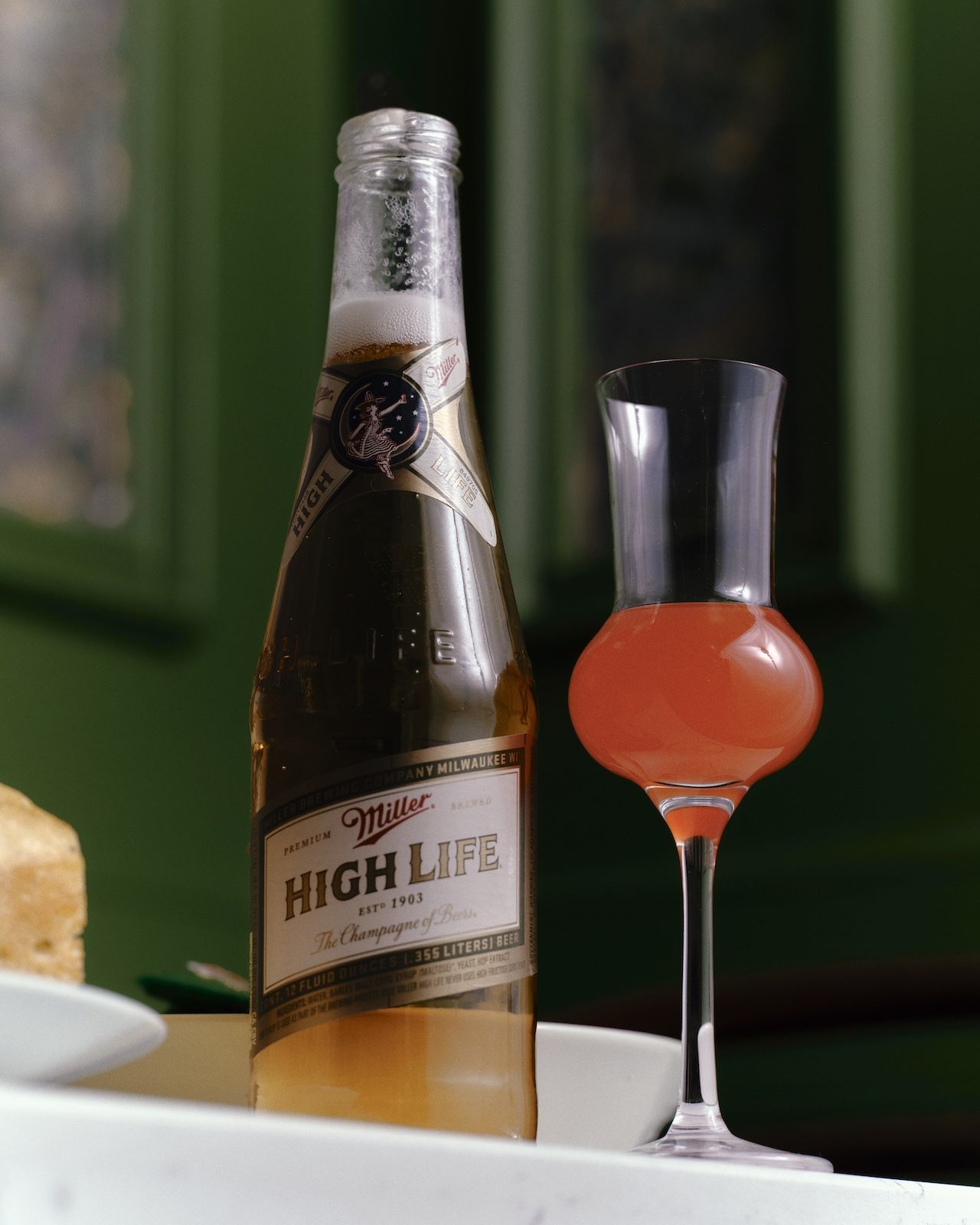 A bottle of Miller High Life beer, with its label reading "The Champagne of Beers," is placed next to a tall, slender glass filled with a pinkish-red drink. The background is a blurred green wall.