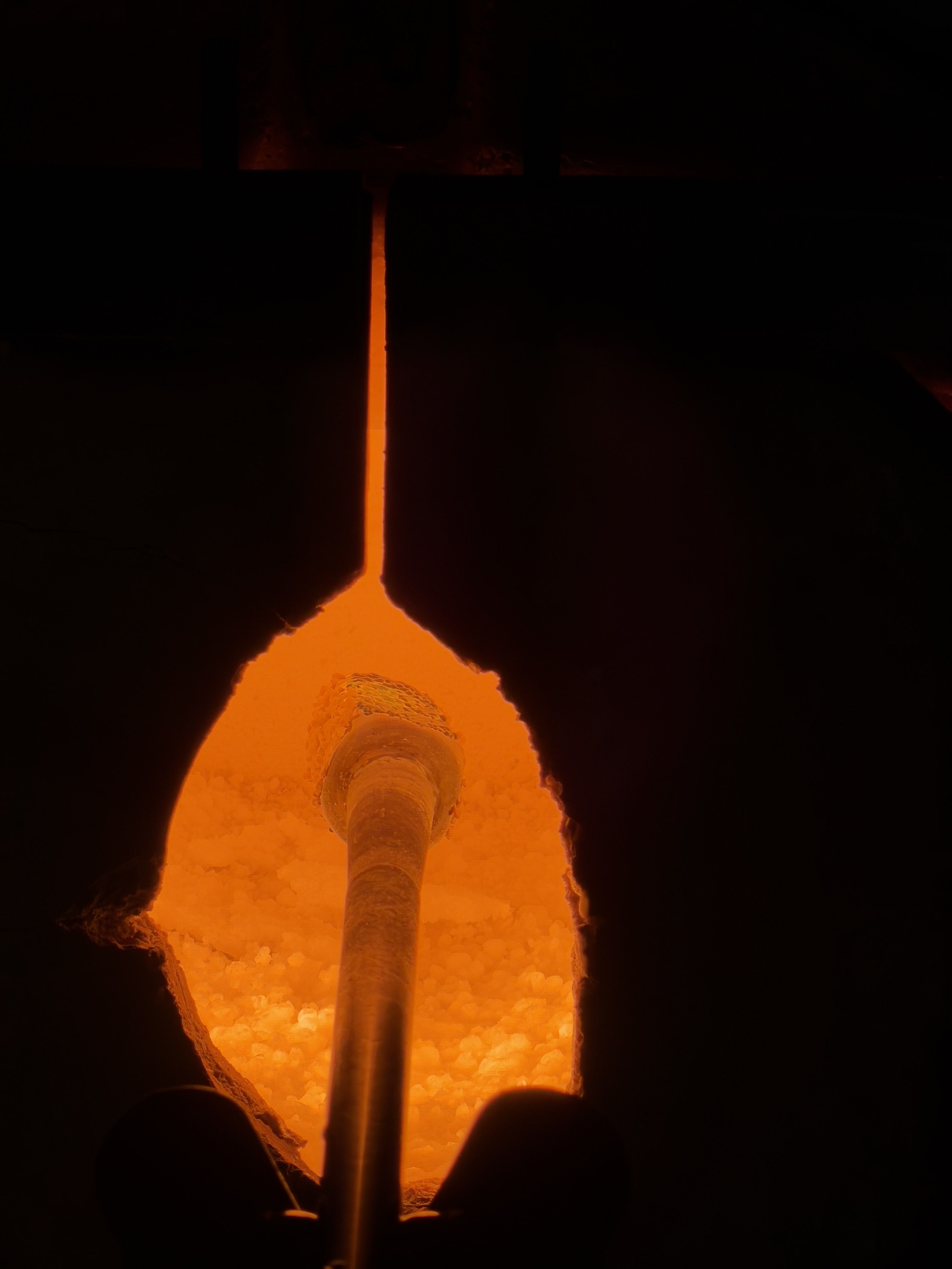 Dark interior of a furnace or lava chamber, bright orange molten material glows at the bottom. A vertical glowing orange pipe rises upward, with dark silhouettes of people in the foreground.