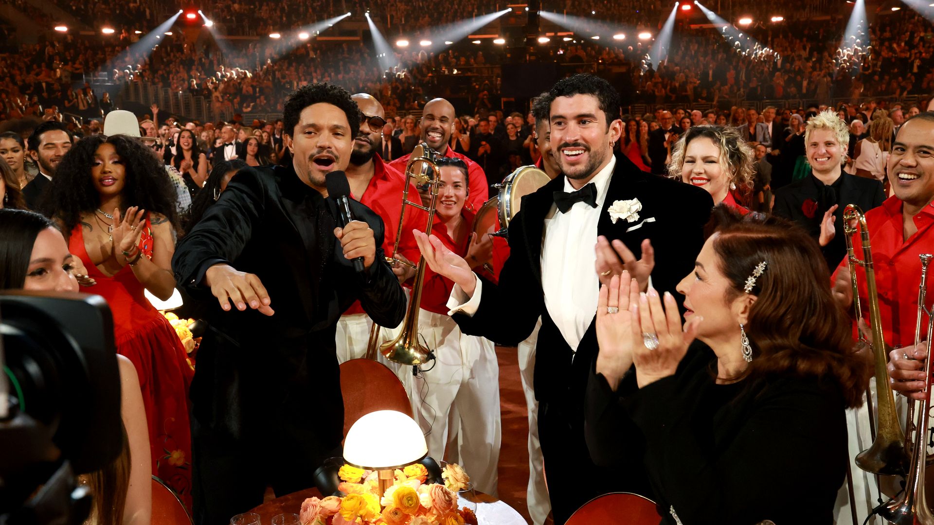 Trevor Noah — wearing all black — and Bad Bunny — wearing a black tuxedo jacket with a white shirt and a black tie — stand together at the Grammy awards with musicians wearing red behind them.