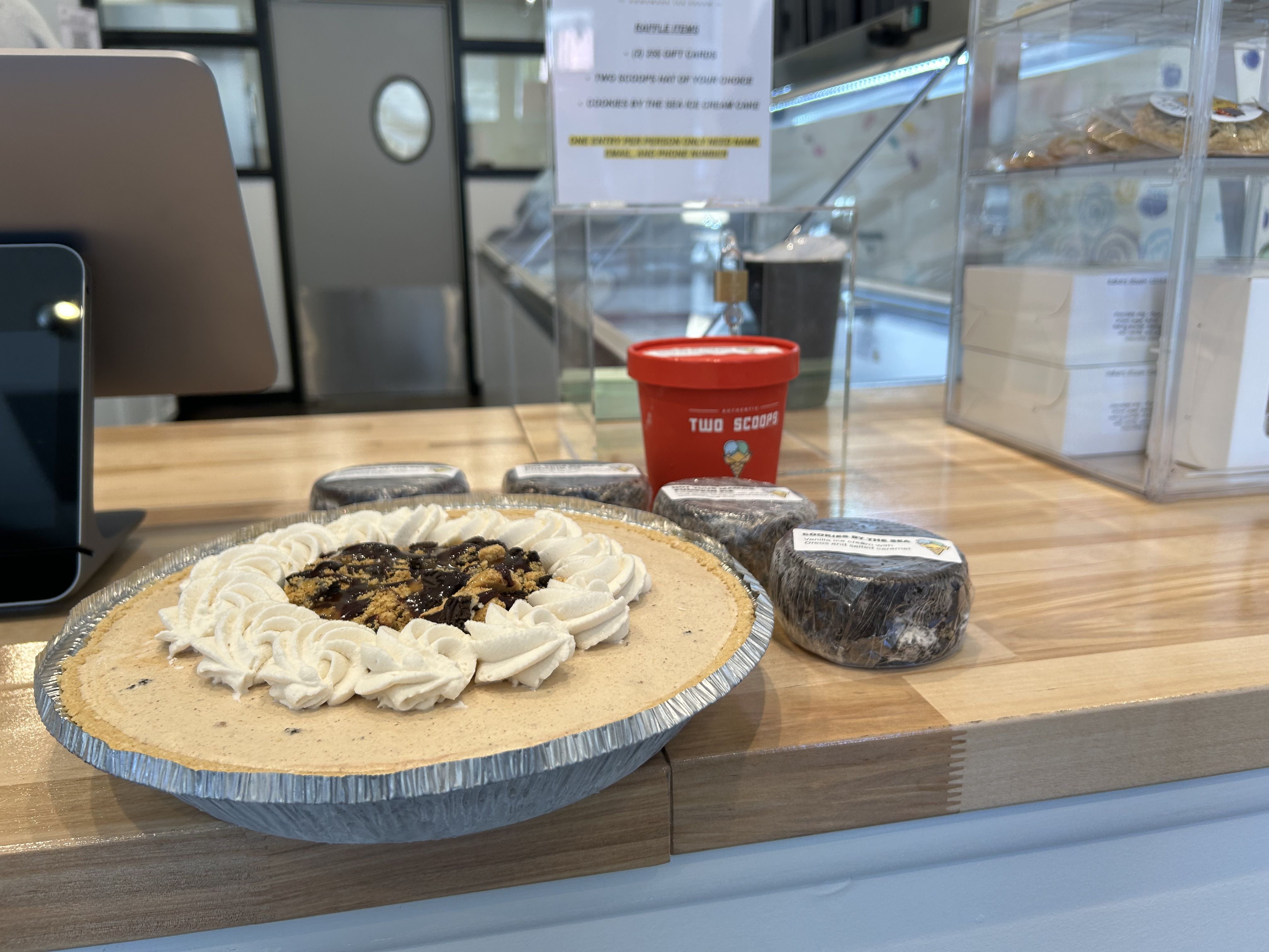 A peanut butter pie with whipped cream and chocolate drizzle on a wooden counter beside wrapped chocolate desserts and a red "Two Scoops" ice cream container in a dessert shop.