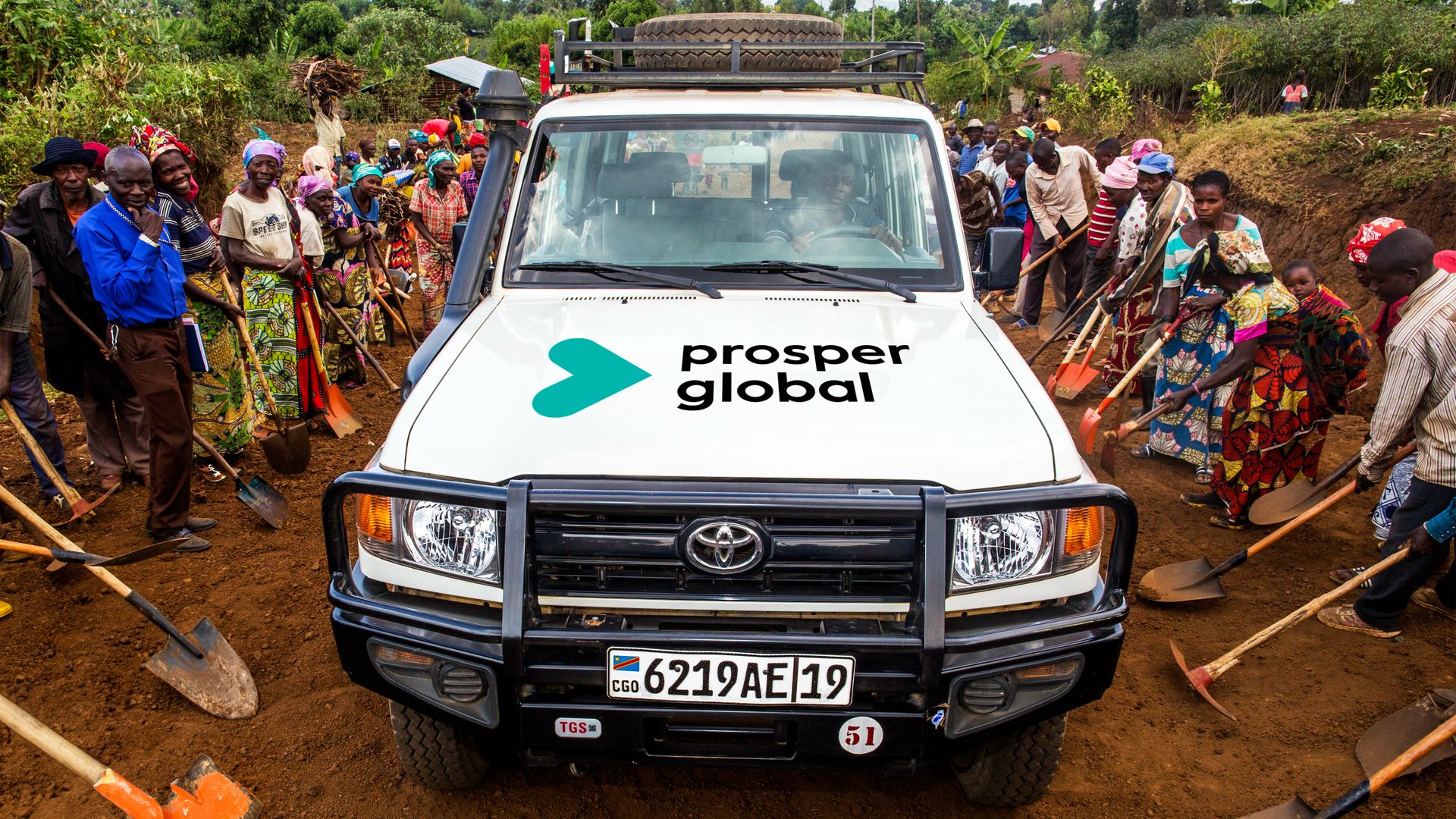 White SUV with the Prosper Global logo on its hood sits on a dirt road, surrounded by a diverse crowd of villagers holding shovels, in colorful clothing; spare tire on the roof.