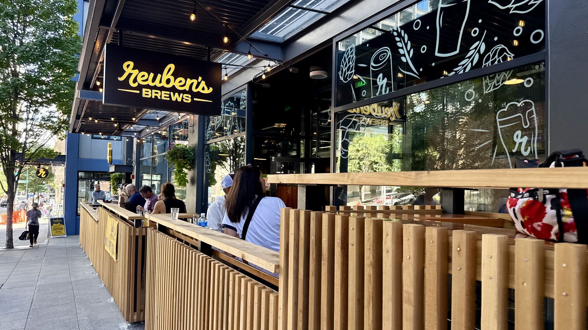 Outdoor seating area at Reuben's Brews with wooden partitions, people seated, string lights overhead, and window illustrations of beer cans and hops in a city street setting.