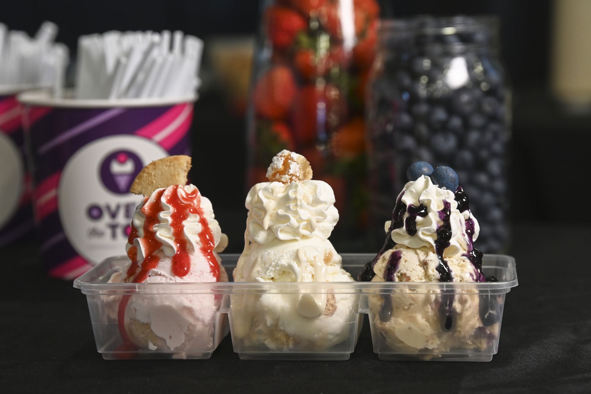 Three clear cups of ice cream with whipped cream toppings: left pink with strawberry sauce and a cookie, middle white with a pastry, right vanilla with blueberries and sauce. Blurred jars and a striped cup sit in the background.