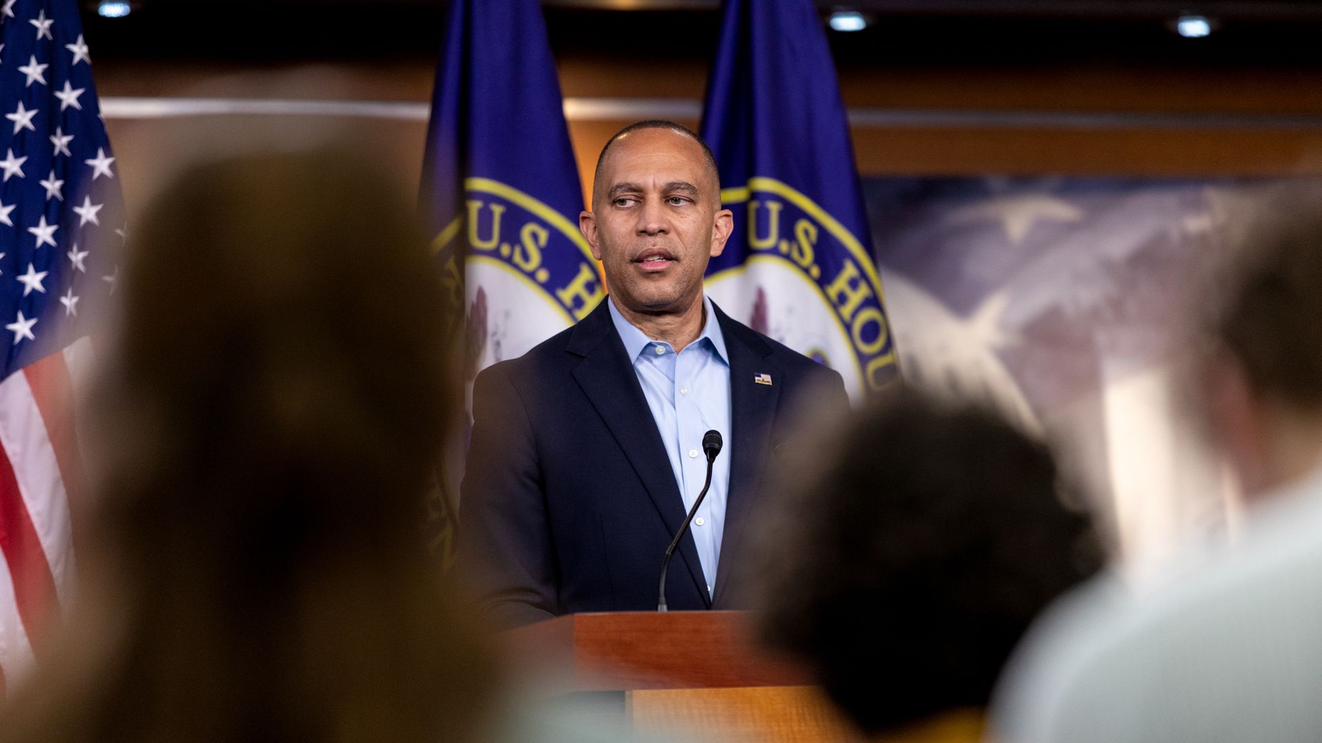 Hakeem Jeffries, wearing a tieless blue suit, speaks at a wooden podium in front of several flags and behind the blurry silhouettes of several reporters.
