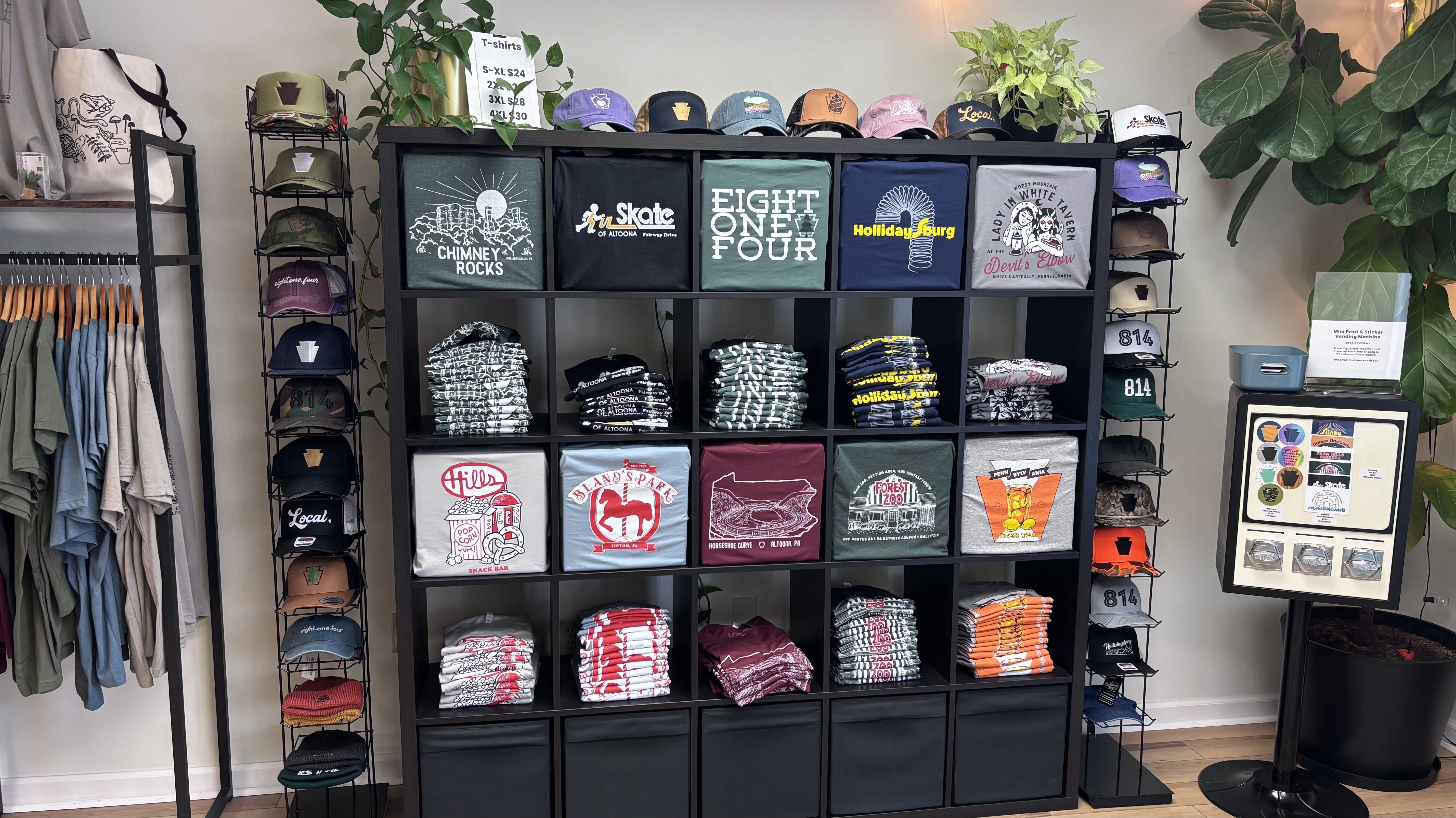 Shop display featuring a black cubby shelf with folded graphic tees and stacked hats. Tops show colorful caps; tees include "Chimney Rocks" and "Eight One Four". Clothing racks and plants frame it.
