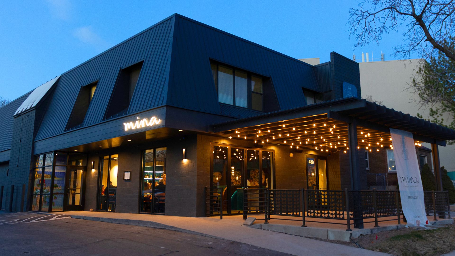 Exterior of a modern restaurant named Mina with a dark blue metal roof. Glass storefront shows diners inside; warm string lights illuminate a covered patio, with a banner and railing at the entrance.