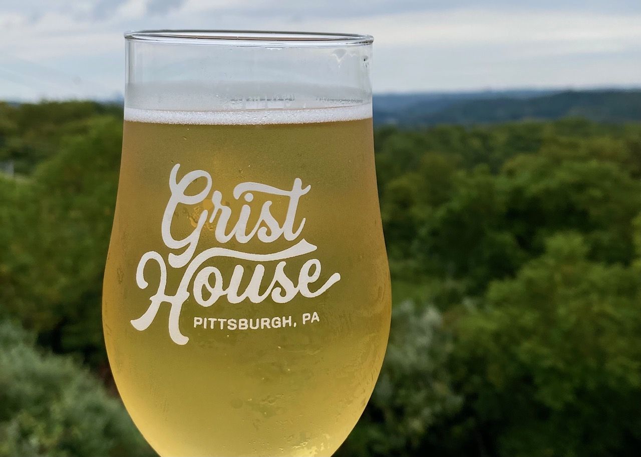 Glass of light golden beer from Grist House Brewing, Pittsburgh, PA, with condensation on glass and blurred green forest and cloudy sky in background.
