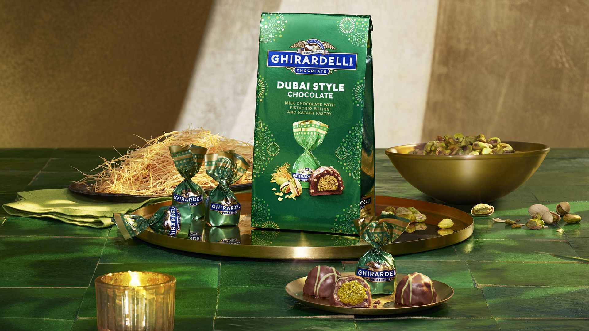 Ghirardelli Dubai Style Chocolate packaging, green wrapped chocolates, pistachios in golden bowl and plate on green tiled surface with warm lighting.