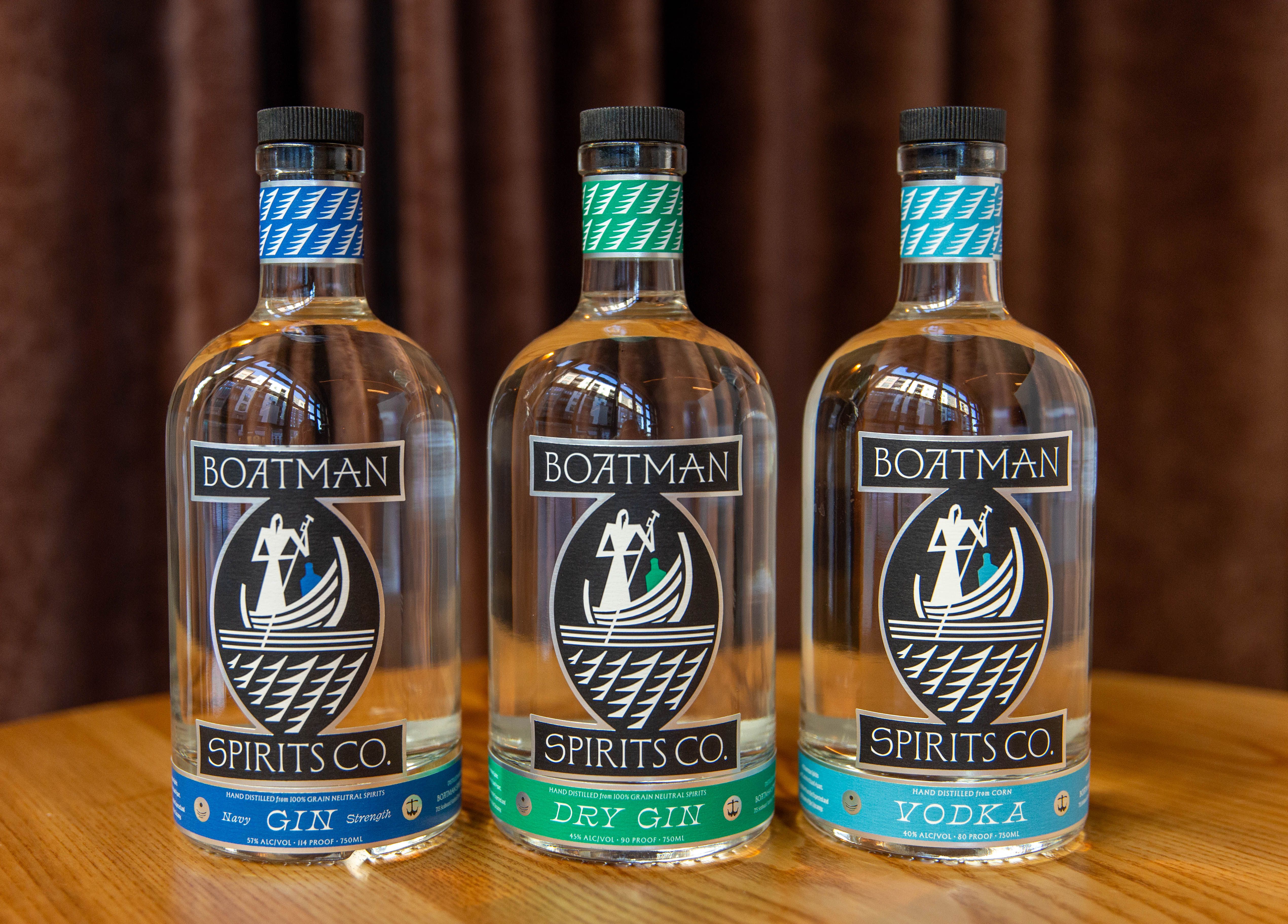 Three clear bottles from Boatman Spirits Co. on a wooden surface with a brown background. Left: Navy Gin, center: Dry Gin, right: Vodka. Each bottle has distinct blue, green, or teal accents.