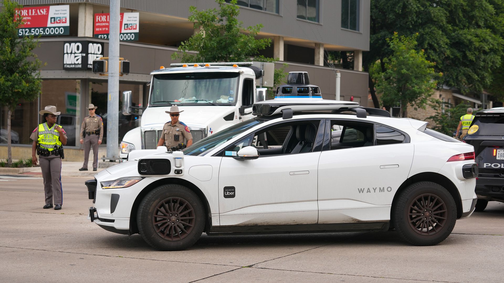 A white Waymo robotaxi stopped at a crash scene, surrounded by police officers, a tow truck and police vehicles. 