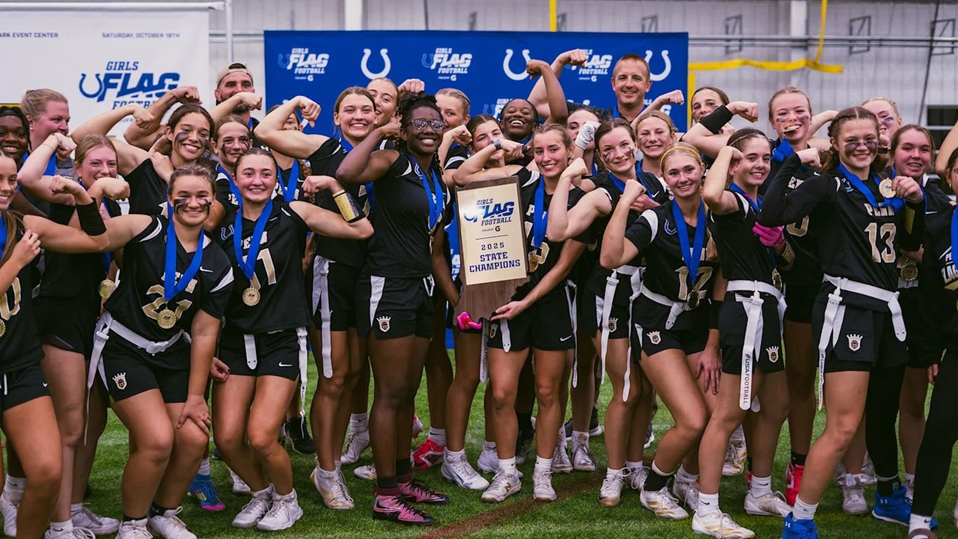 Girls flag football team in black uniforms with medals pose on a field, fists raised, holding a state champions plaque against a blue backdrop.