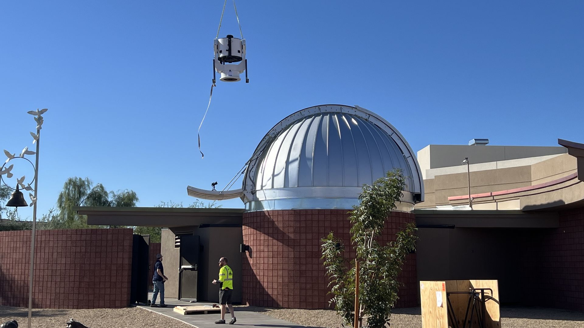 Silver-domed observatory building with red brick walls under clear blue sky; telescope equipment being lifted by crane; two people near the entrance; small tree and bird sculpture nearby.