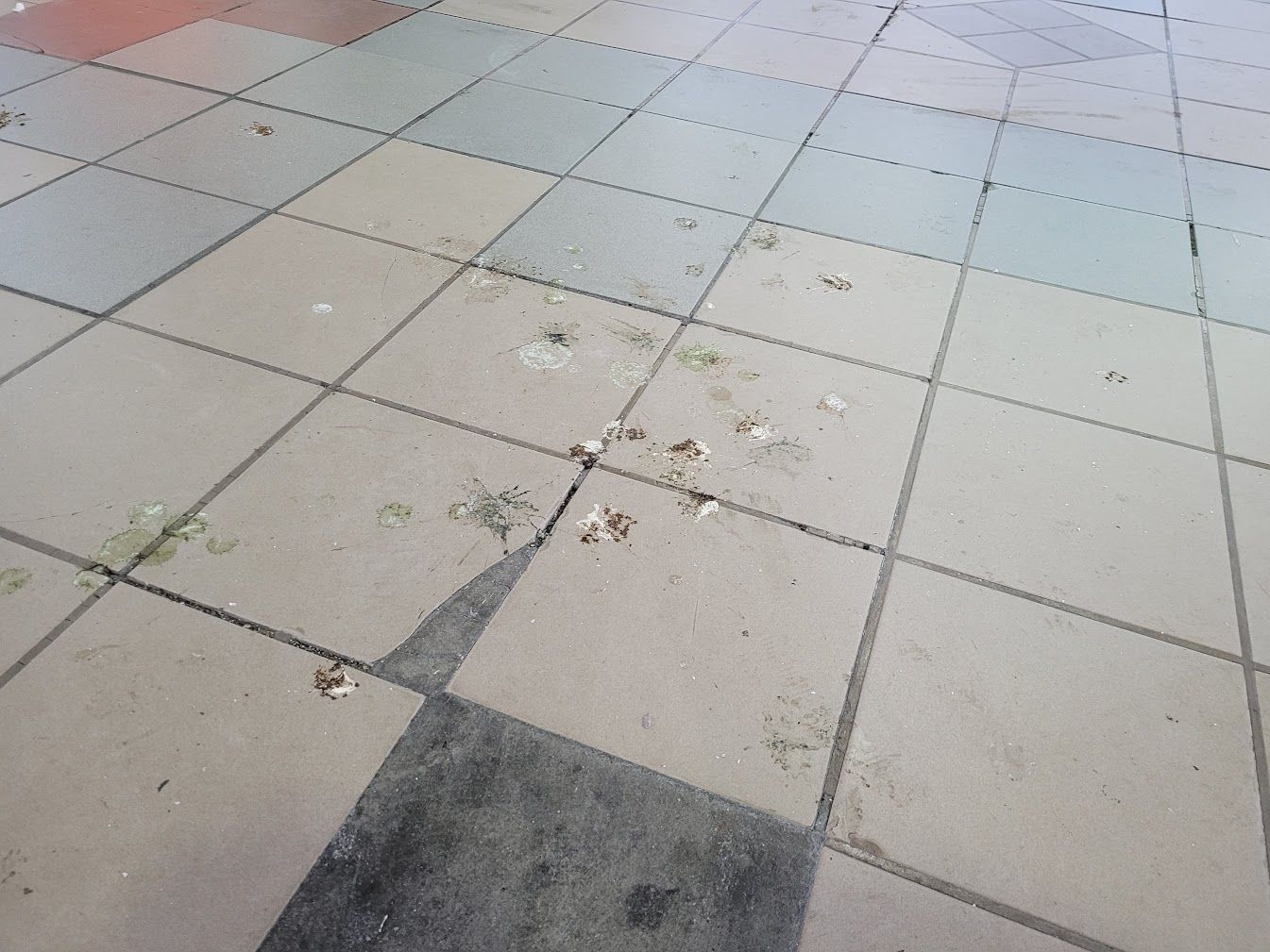 Wide view of a tiled pavement with pastel beige, gray, and blue squares, showing dirt, scuffs, and small footprints scattered across the surface.