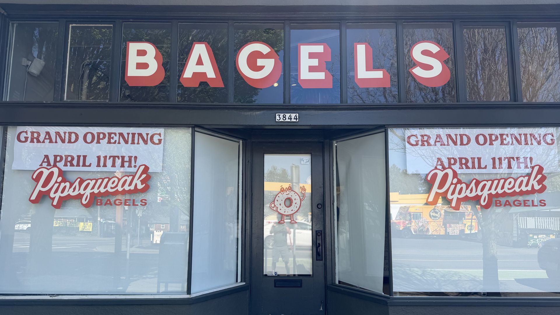 Storefront with large red BAGELS letters on front windows; banners announce "GRAND OPENING APRIL 11TH!" and "Pipsqueak BAGELS". Center entry door shows a small donut sticker, street reflected.