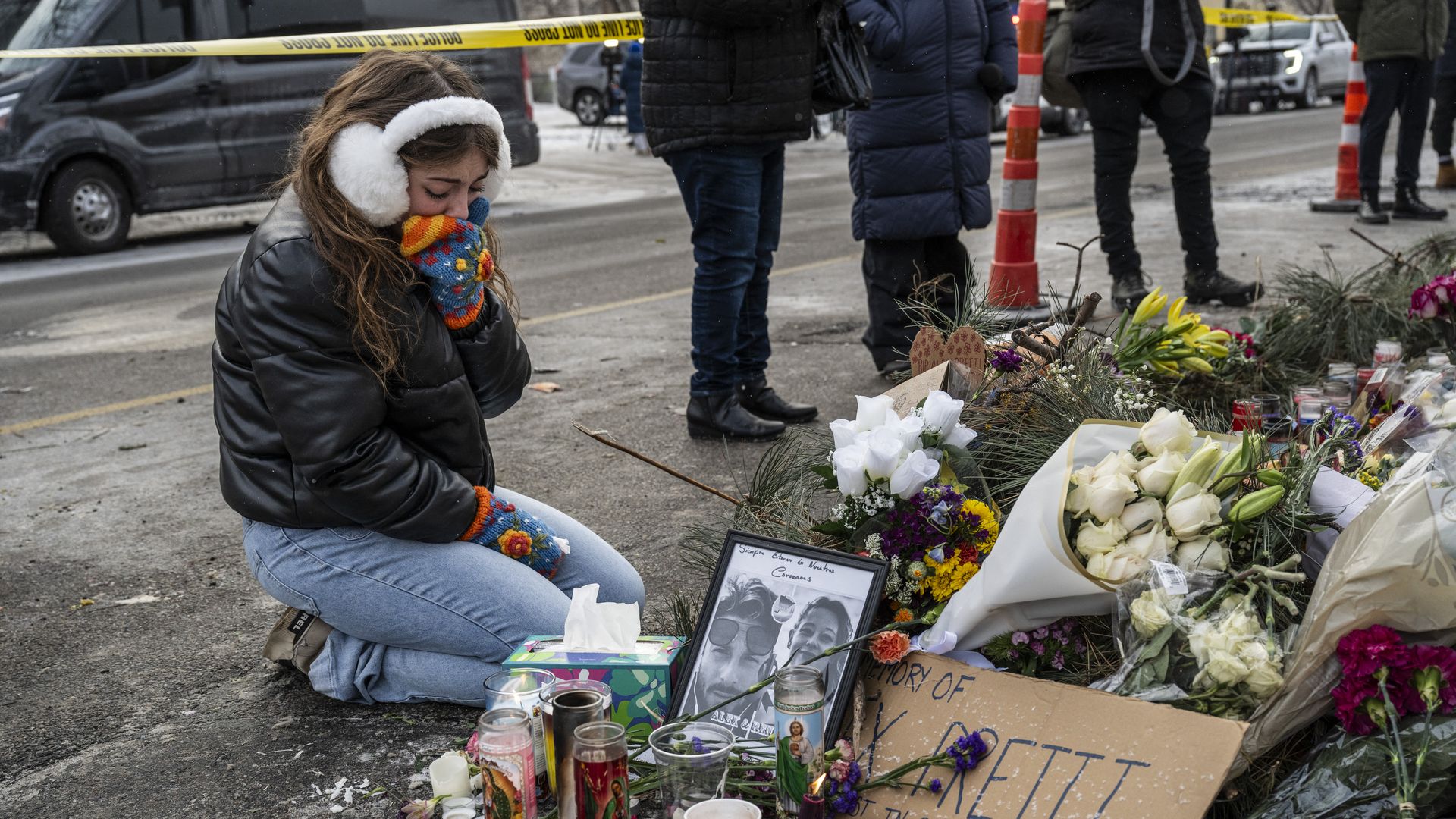 A woman wearing white earmuffs and colorful mittens kneels at a memorial for Alex Pretti made of candles and flowers.