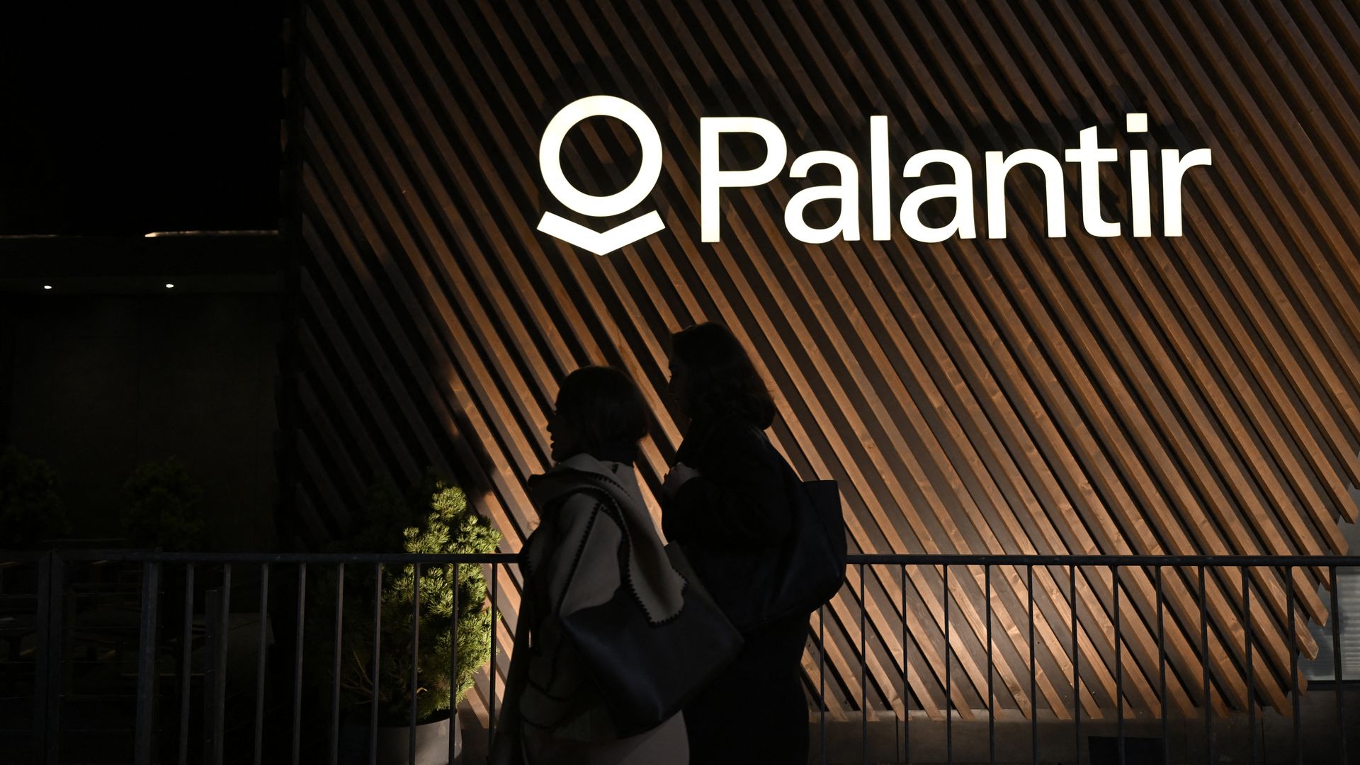 Silhouettes of two people walking past a wooden slatted wall with the illuminated white Palantir logo at night.