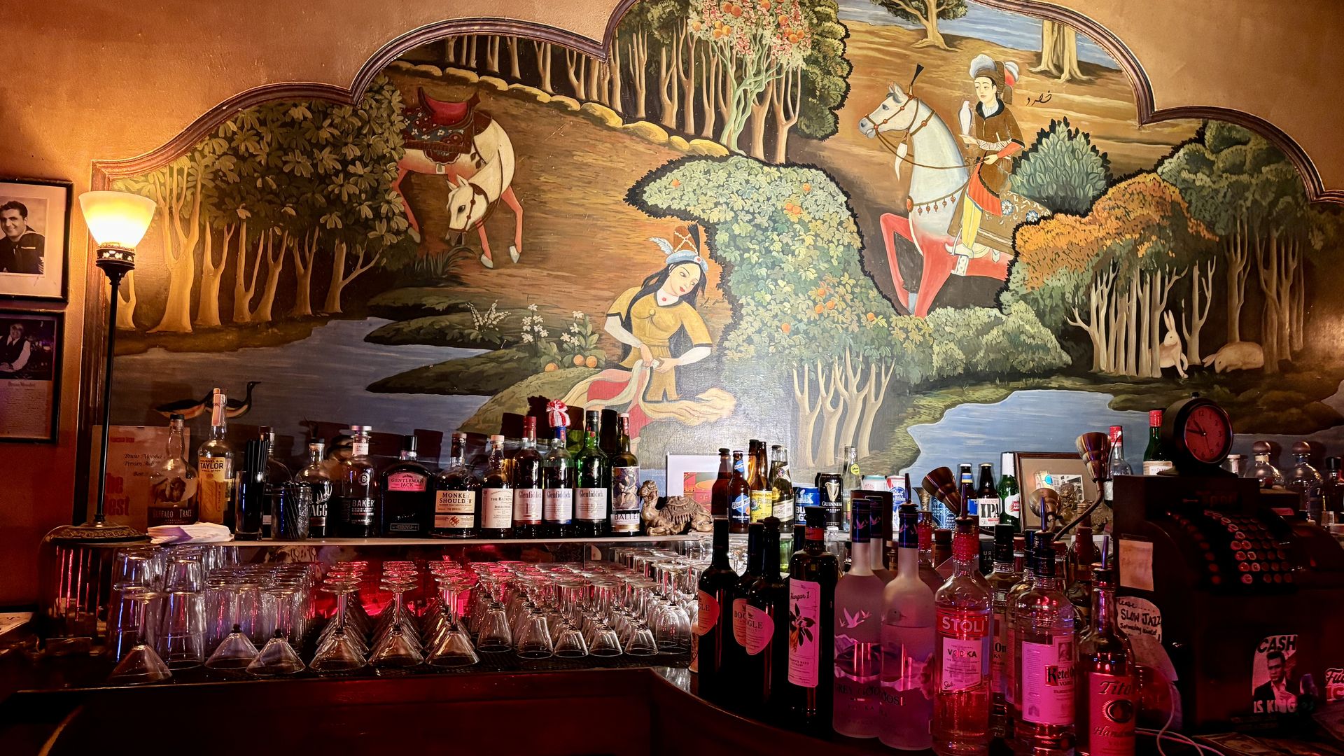 Warm bar scene with bottles and glassware in the foreground. A large mural shows a woman gathering fruit by a river, and riders on horseback among trees, creating a fantasy pastoral backdrop.
