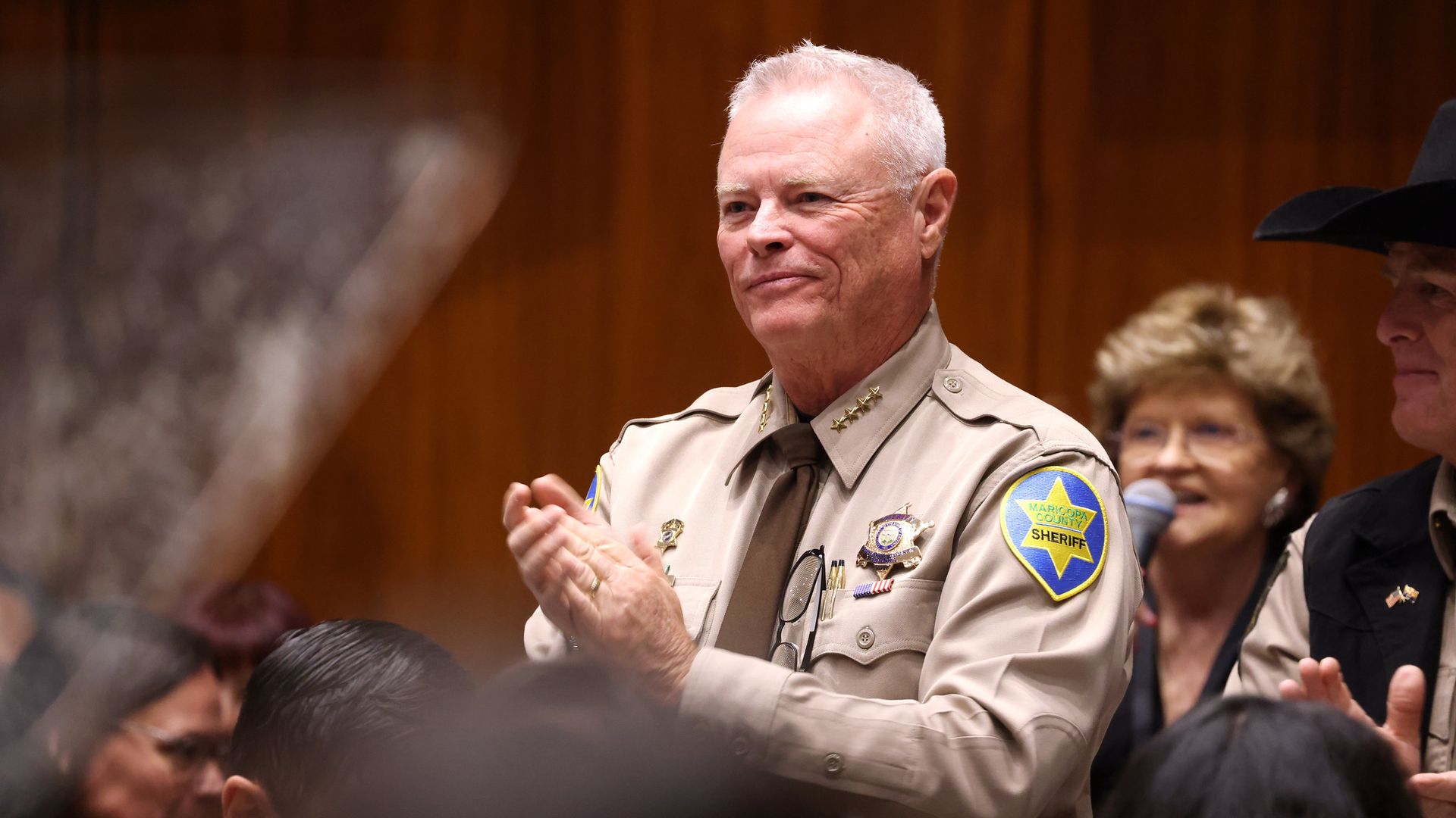 Smiling Sheriff Jerry Sheridan in a tan uniform with badges and a sheriff patch stands in a crowd at an indoor event, with a wood-paneled backdrop behind.