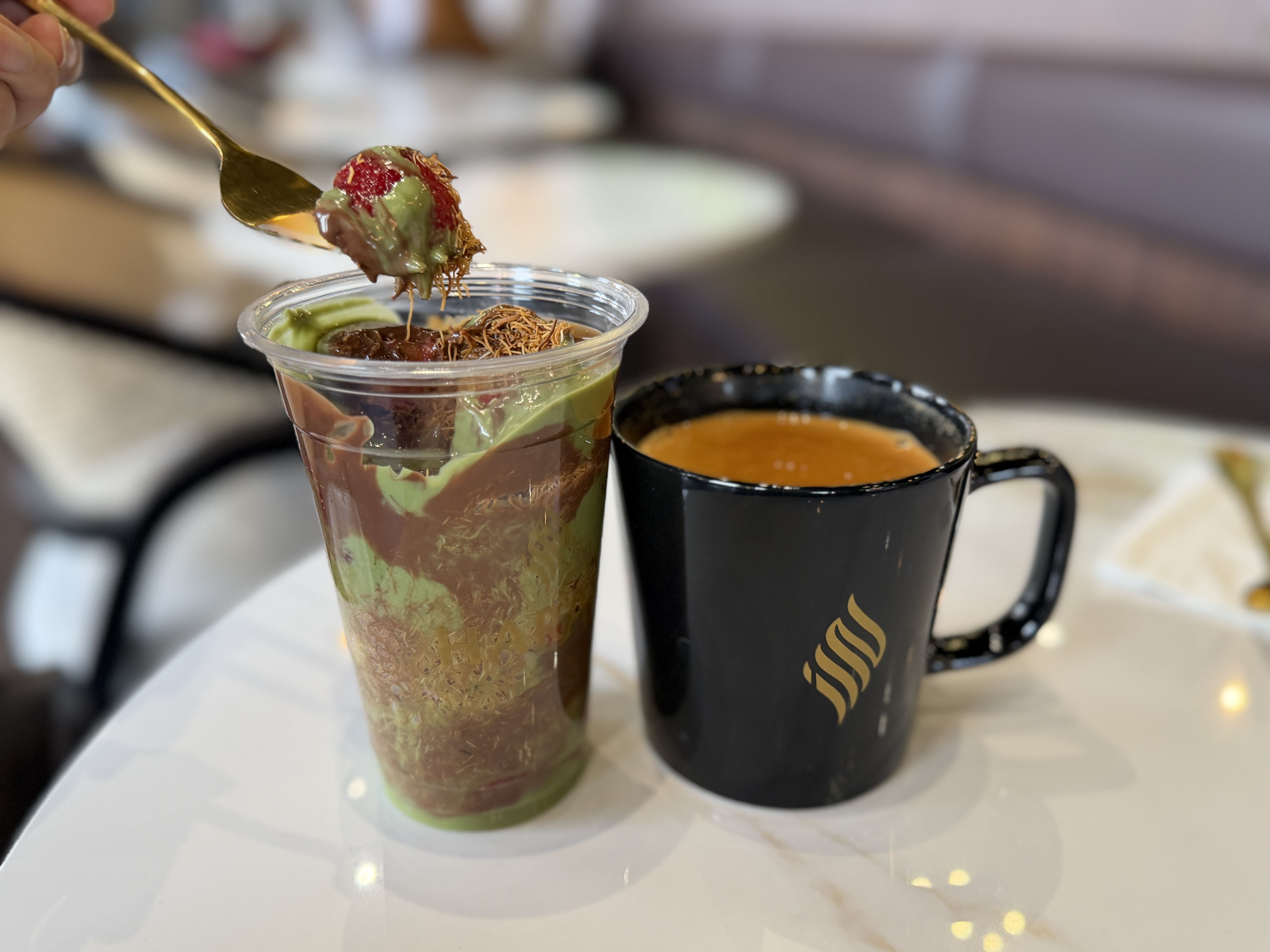 A spoon lifts a bite of a layered green and brown dessert from a clear cup, topped with shredded garnish and a red berry; a black mug of orange tea sits on a white table in a cafe.