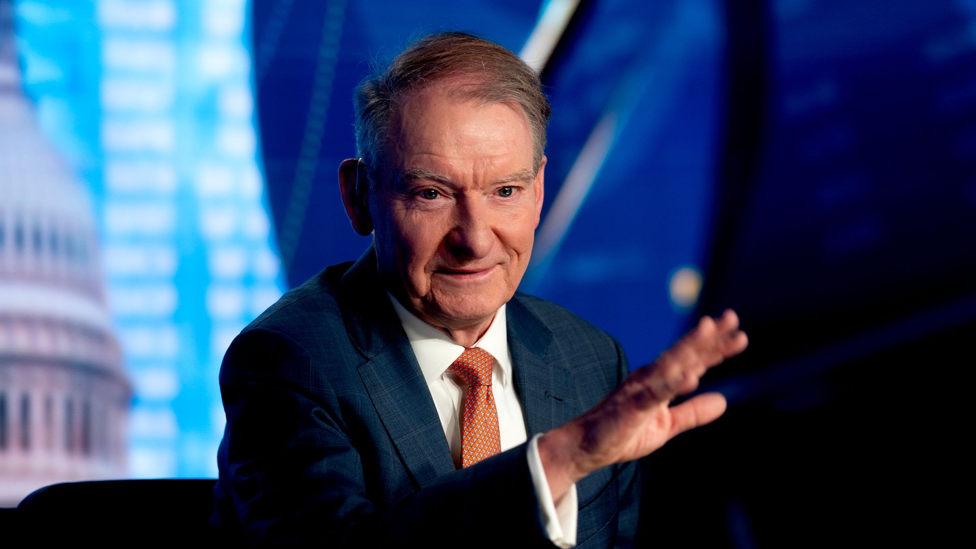 Older man in a dark suit and orange tie gesturing with his hand, seated against a backdrop featuring a blurry U.S. Capitol dome and blue digital patterns.