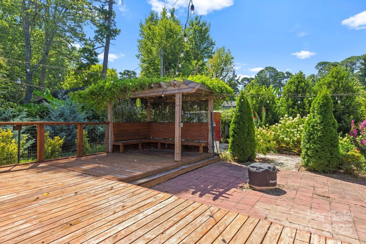 Wooden deck with built-in bench seating under a pergola covered with green vines, surrounded by string lights, shrubs, and tall trees under a bright blue sky.