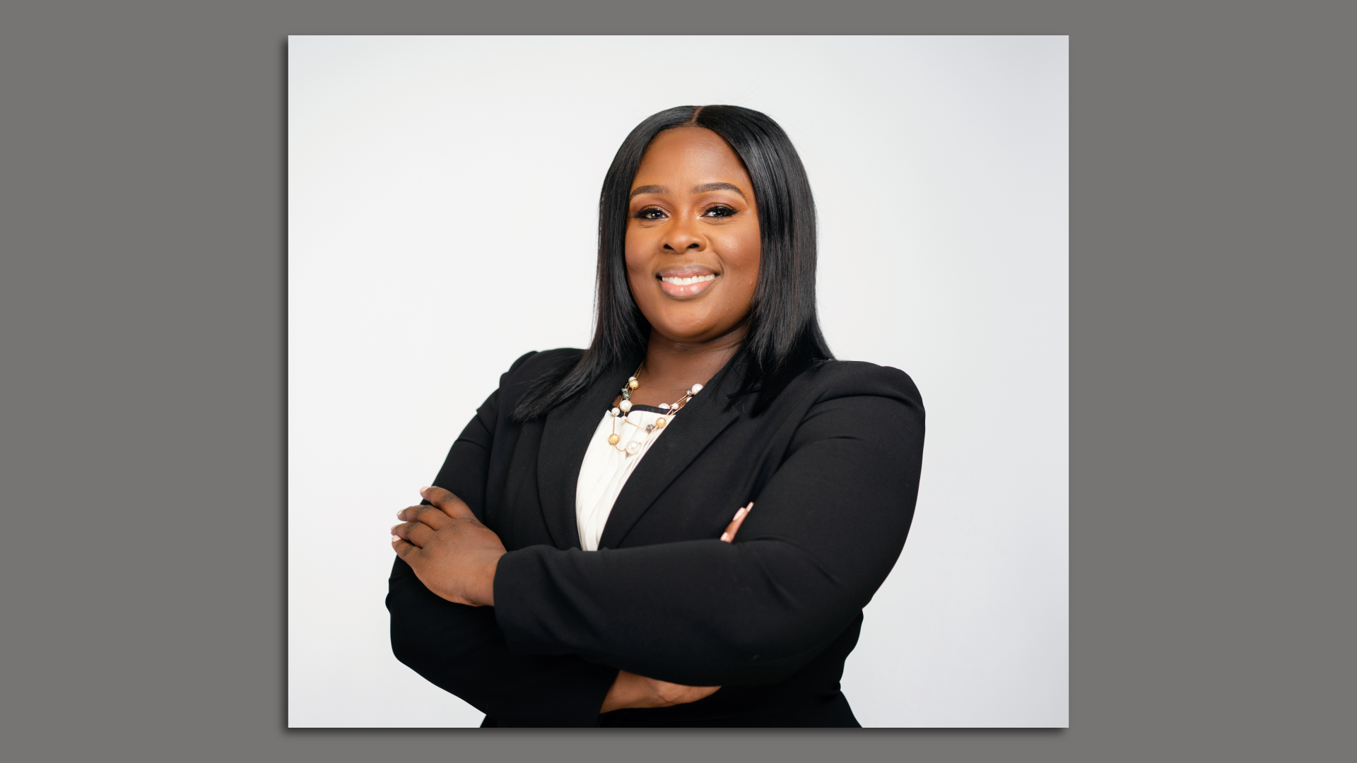 An individual wearing a black pantsuit smiles while folding their arms in front of a white background